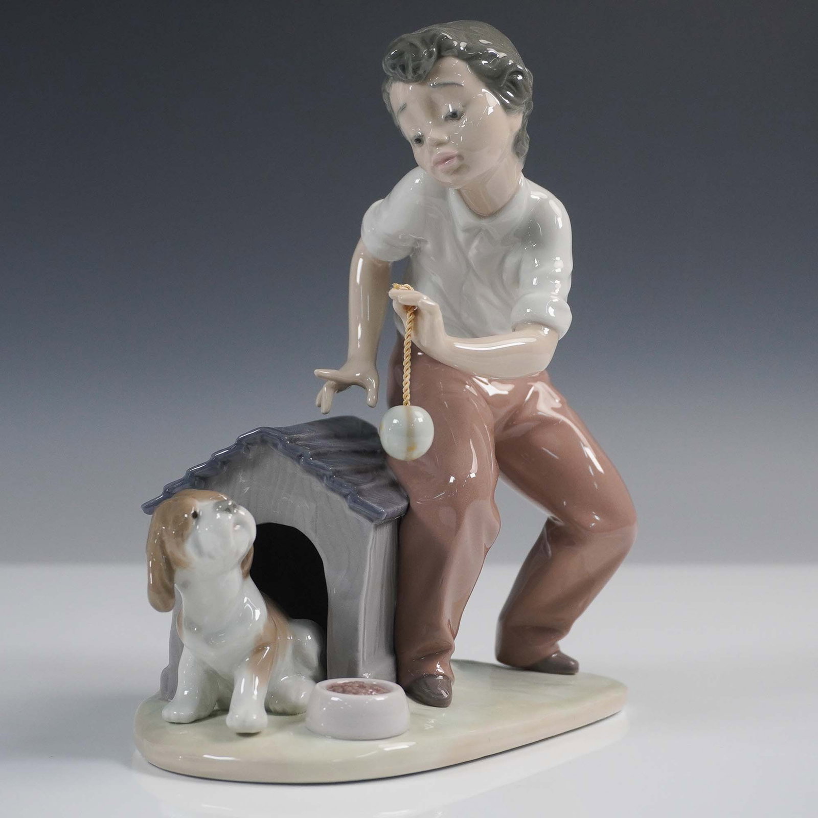 Lladro Porcelain Figurine, Come Out and Play 1005797: Charming porcelain figure in a glazed finish depicts a tender moment of a young boy playing ball with his young puppy. Lladro backstamp. This item has its original box. Box measures: 10"L x 9"W x 6.25