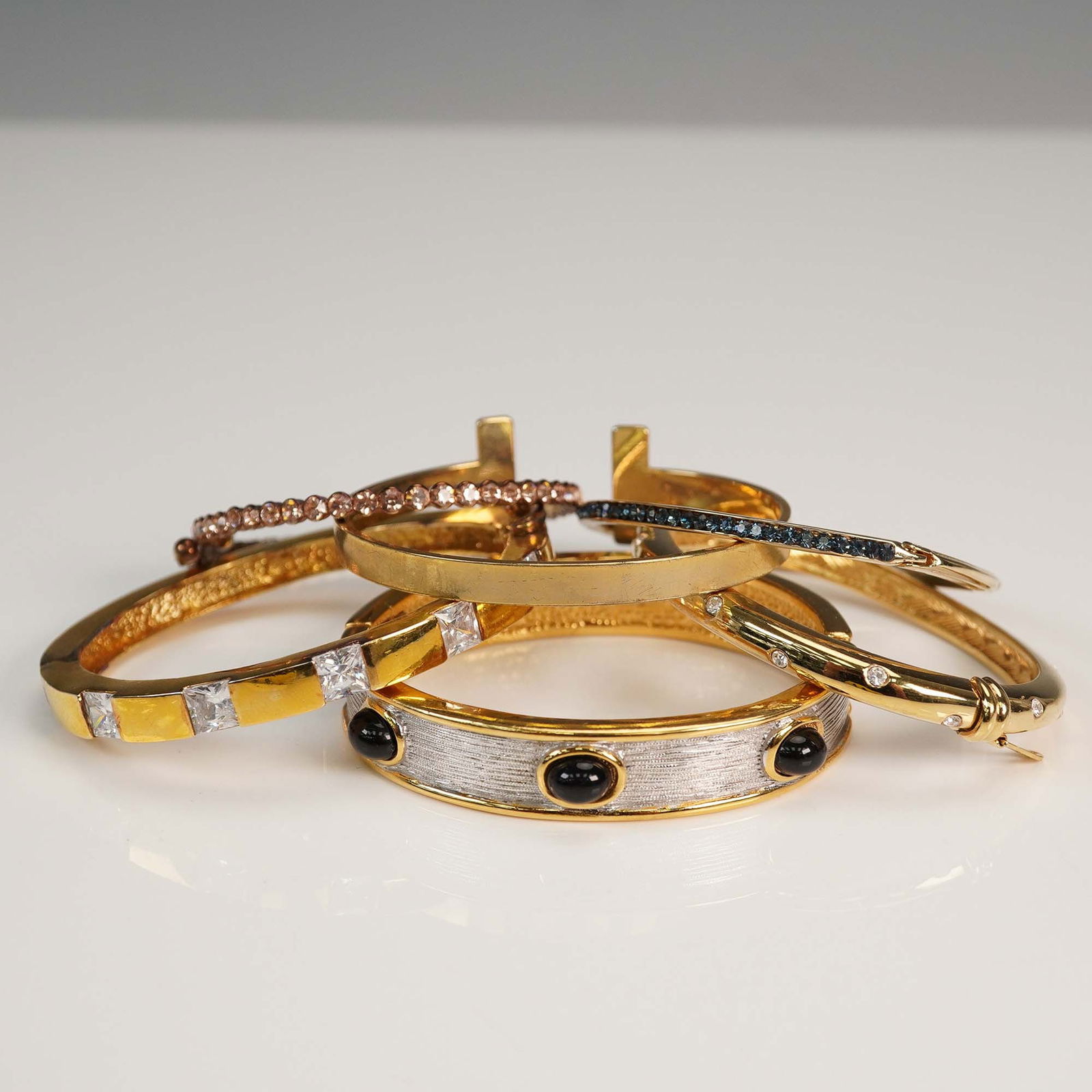 6pc Gold Tone Hinged Bangles: Collection of six gold tone hinged bangles featuring a variety of decorative styles. Designs include clear square-cut and round-cut rhinestones, bezel-set black cabochons on a textured silver-tone pan