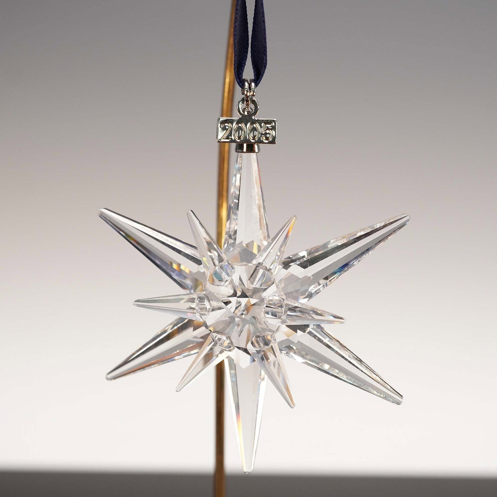 Swarovski Crystal 2005 Christmas Ornament: An exquisite Swarovski Crystal 2005 Christmas Ornament, featuring a stunning clear crystal snowflake design suspended from a white satin ribbon. The ornament, meticulously crafted in Austria, is etche