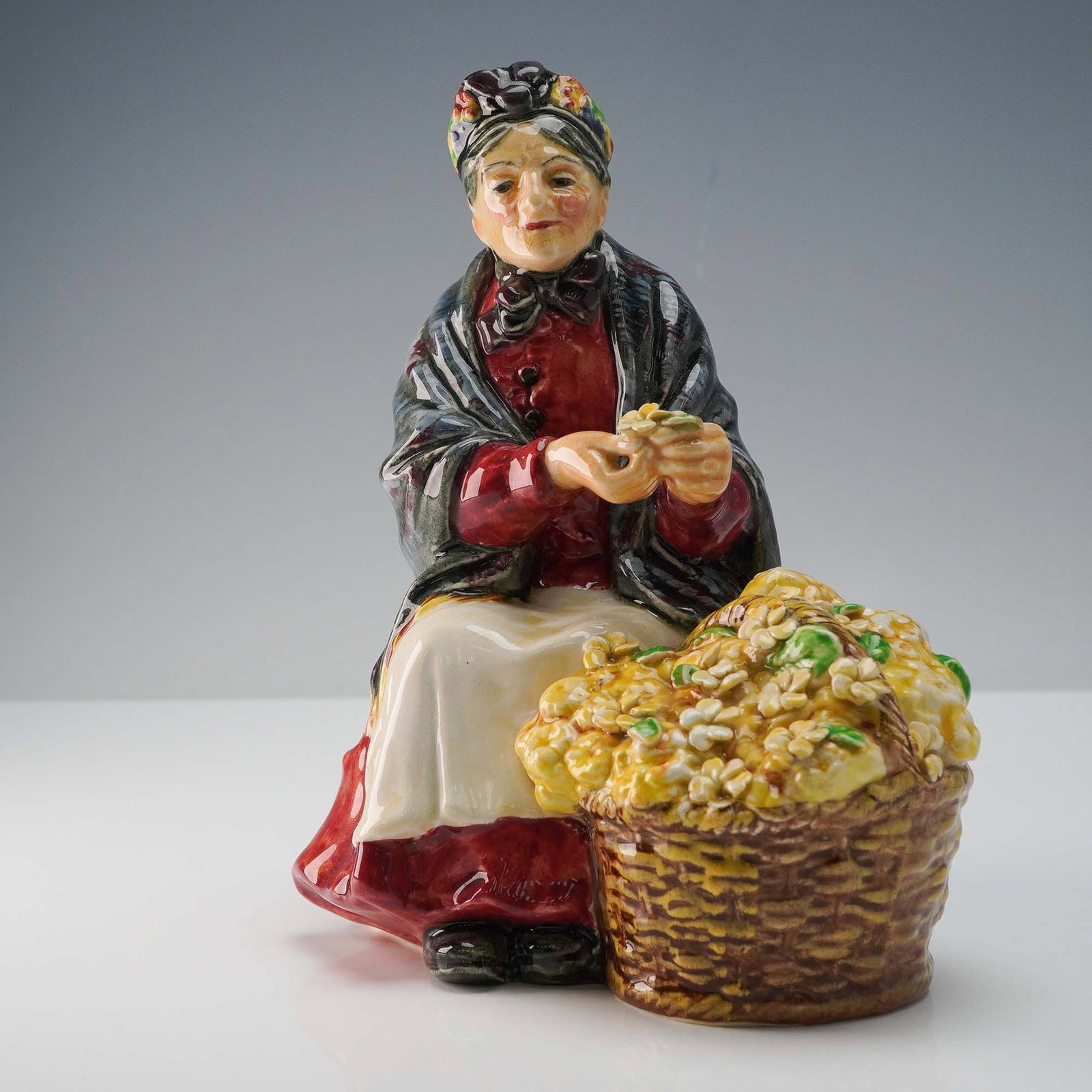 Royal Doulton Figurine, Primroses HN1617: Primroses HN1617 by Royal Doulton features an elderly flower seller seated beside a large basket overflowing with yellow blossoms. Dressed in a red skirt and floral bonnet, she holds a small handful o