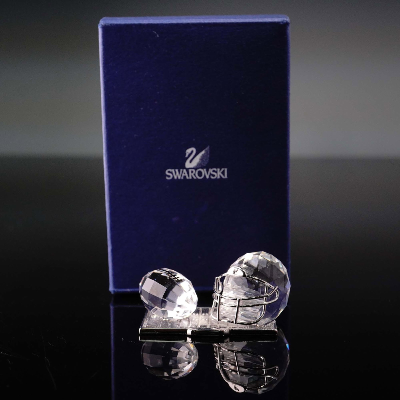 Swarovski Crystal Figurine, American Football Trophy 695804: This retired Swarovski Crystal Memories American Football Trophy figurine showcases a faceted clear crystal football and helmet on a rhodium-plated base. Part of the Crystal Memories Sports and Trophi