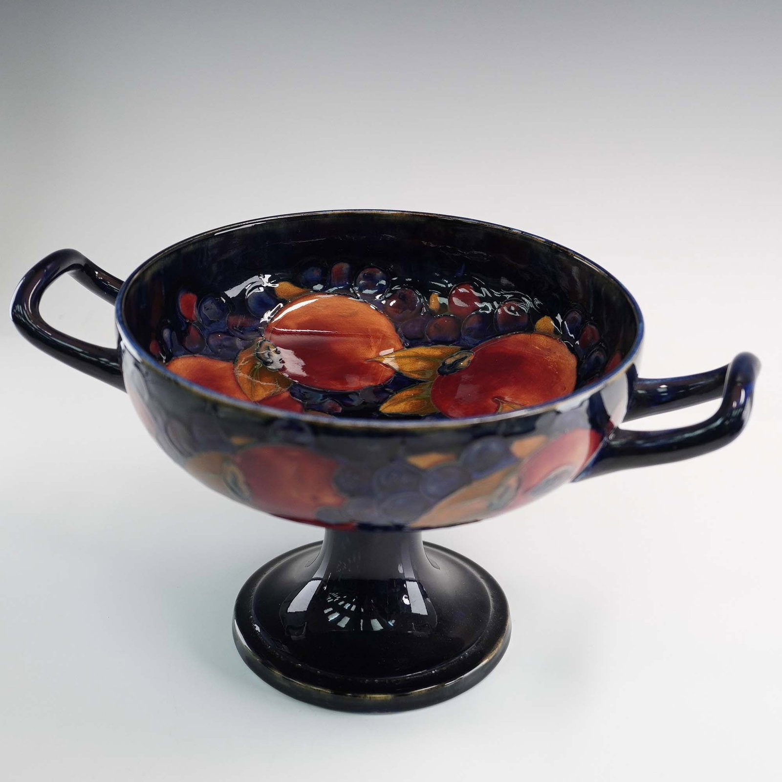 Moorcroft Pottery Pedestal Comport Pomegranate Pattern: This rare pedestal comport by William Moorcroft features the celebrated Pomegranate pattern, with richly glazed fruits and berries rendered in deep crimson, ochre, and violet tones against a flowing c