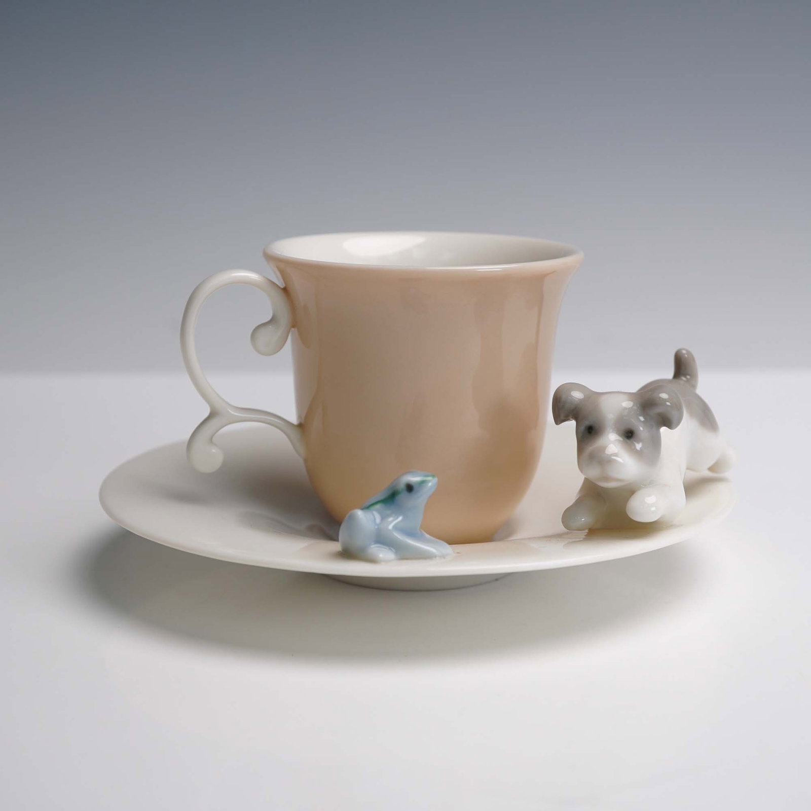 Lladro Porcelain Cup and Saucer Set, Cautious Friends 1006046: A playful porcelain cup and saucer set pairs a peach hued cup with a white saucer, featuring a curious dog and a bright blue frog figurine perched on the saucer' edge. These whimsical accents bring a 