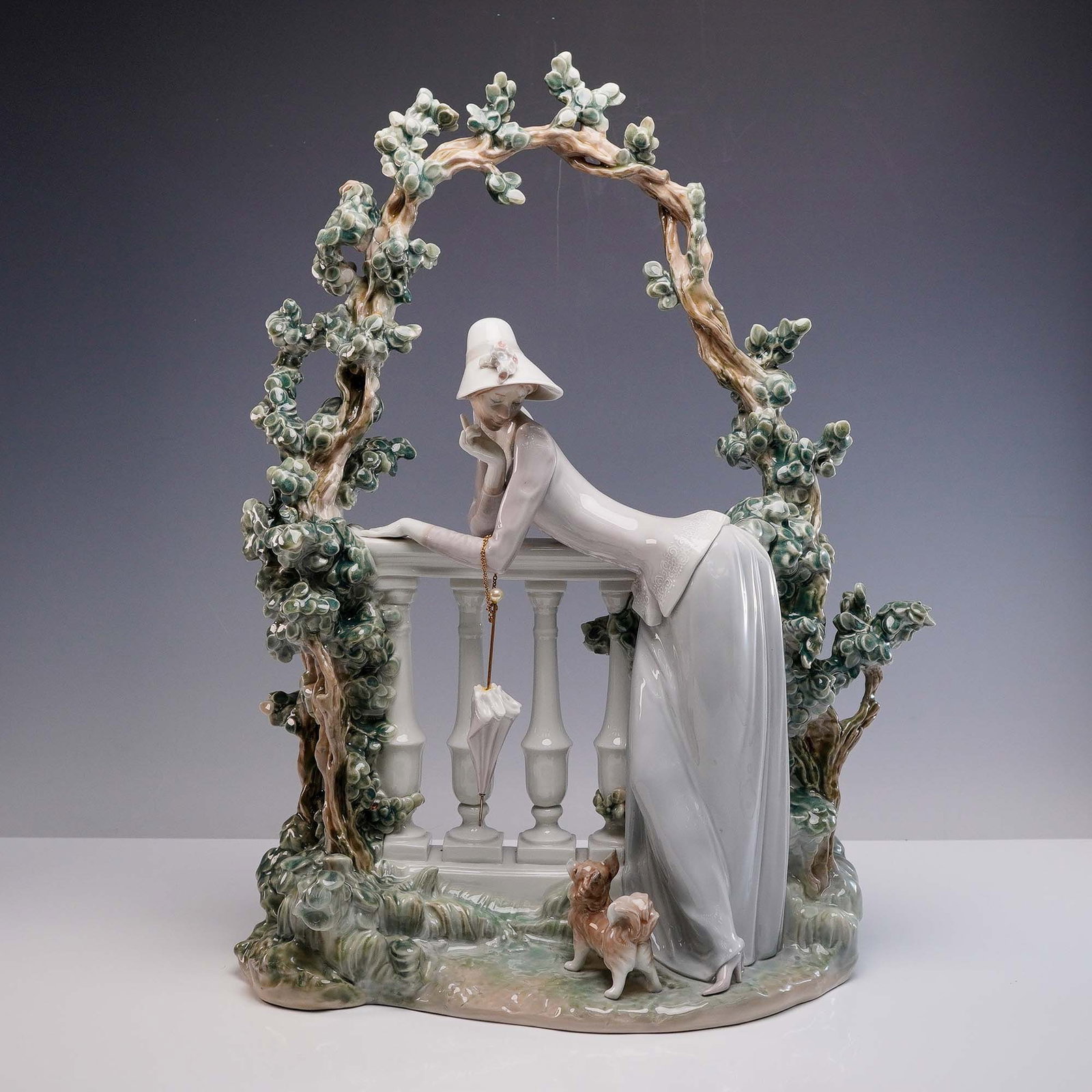 Lladro Porcelain Sculpture, Thoughts 1001272: Glazed finish porcelain sculpture depicting a graceful lady in a flowing gown and wide brim hat, resting thoughtfully against a balustrade beneath a floral arch, with a small attentive dog at her feet