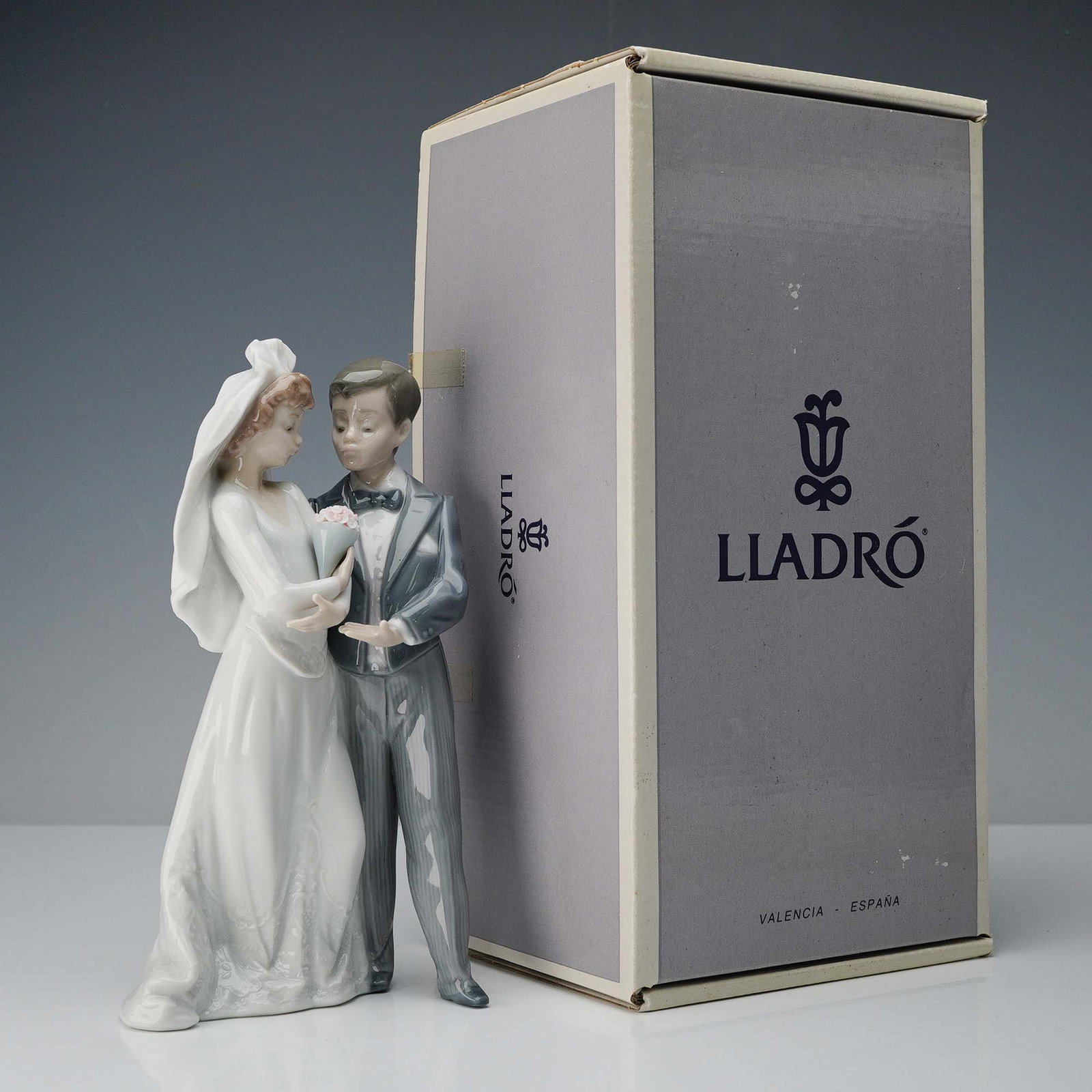 Lladro Porcelain Figurine, From This Day Forward 1005885: Depicting a bride and groom on their wedding day, the figurine is rendered in Lladro's signature glossy finish. The bride is elegantly dressed in a long, flowing gown with a veil and holds a small bou