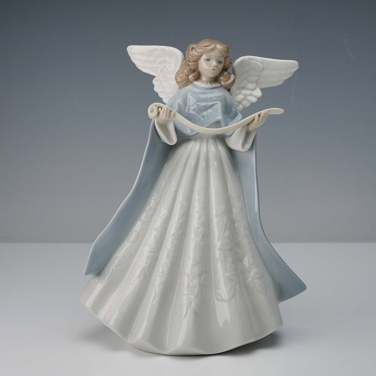 Lladro Porcelain Figurine, Tree Topper '90 - Blue 1005719: Depicts a serene angel holding a music scroll. Rendered in a glossy finish, the figure is adorned with a flowing white gown featuring delicate floral patterns and a soft blue cloak that extends gracef