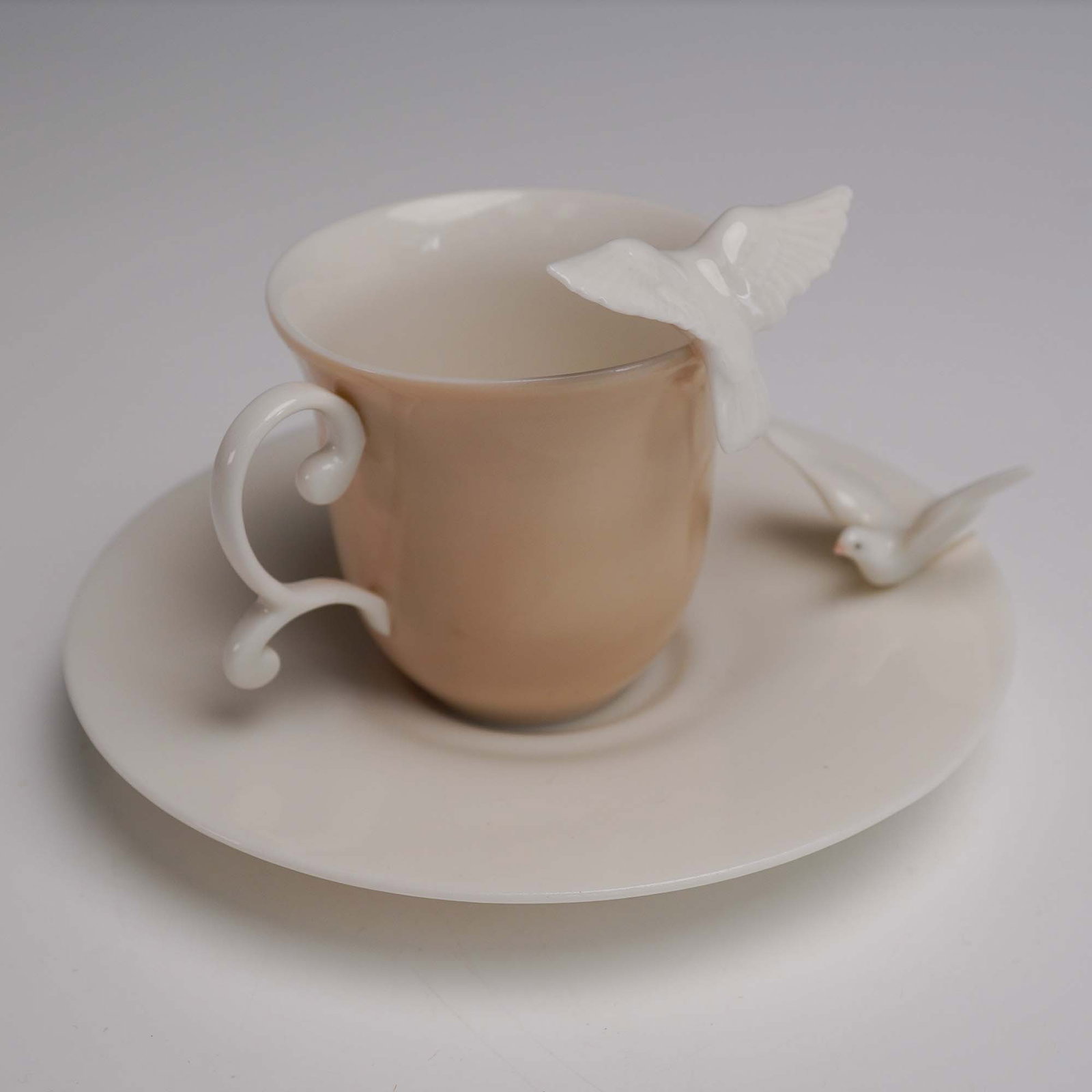 Lladro Porcelain Cup and Saucer Set, Doves 1006043: Elegant porcelain cup and saucer set features a soft peach colored cup and a creamy white saucer, enhanced by delicate white dove figurines perched around the rim and saucer edge. The cup's handle fea