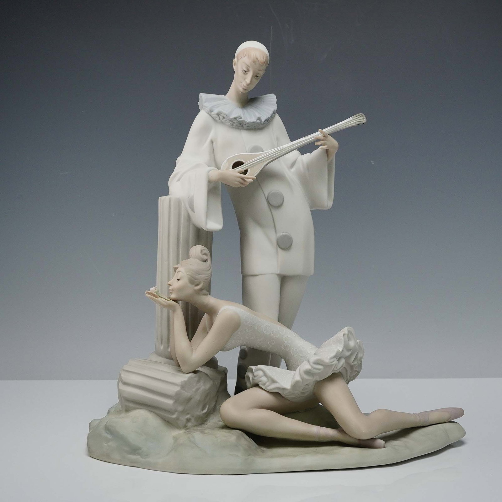 Lladro Porcelain Sculpture, Pensive Pierrot 1014560: This elegant porcelain figurine captures a poetic moment between a delicate ballerina and the wistful figure of Pierrot, who gently strums a lute beside a classical column. Rendered in soft matte glaz