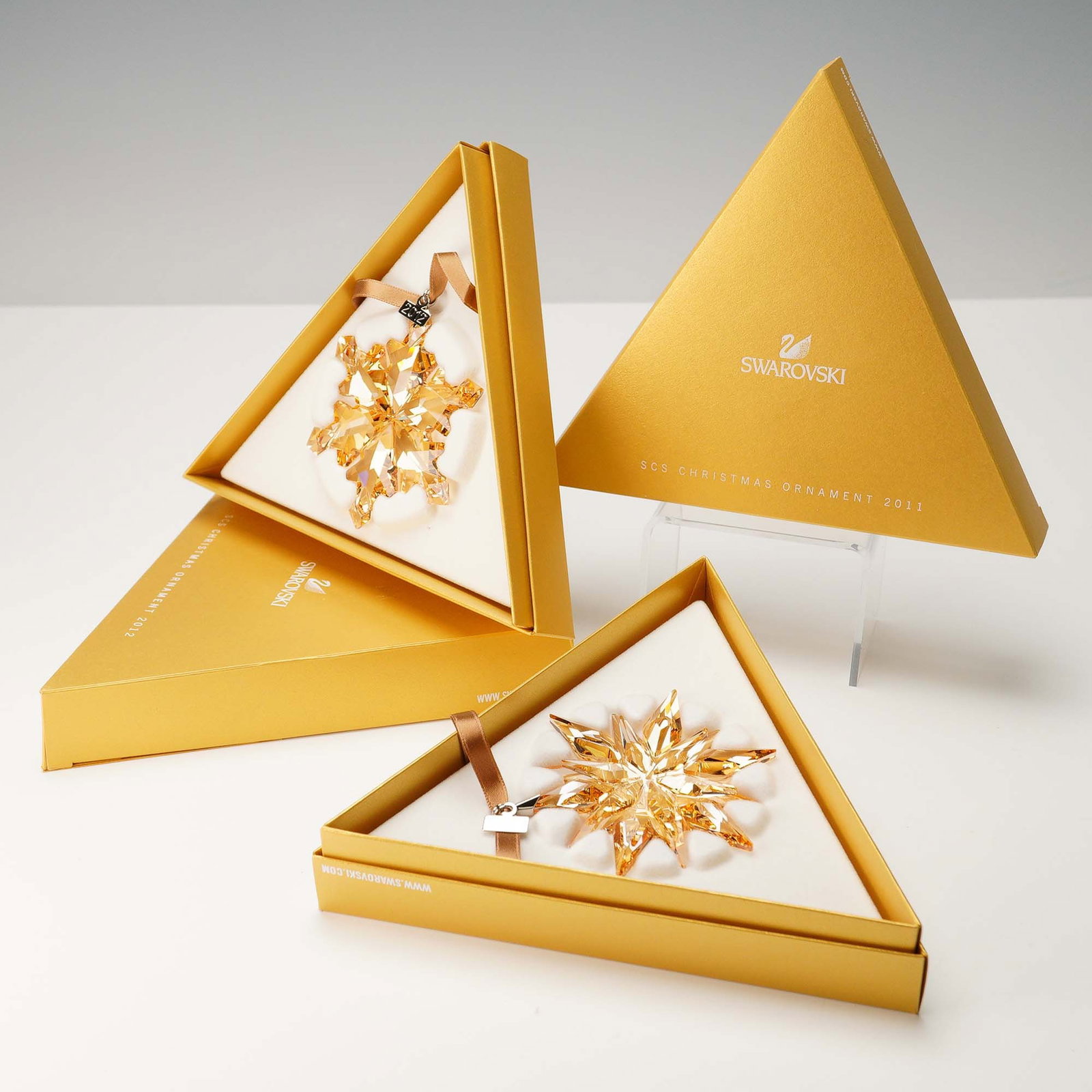 2pc Swarovski SCS 2011 and 2012 Gold Christmas Ornaments: Pair of Swarovski Crystal Society (SCS) Christmas ornaments from 2011 and 2012. Both ornaments are designed in the shape of snowflakes and crafted from golden-shaded faceted crystal. Each is suspended