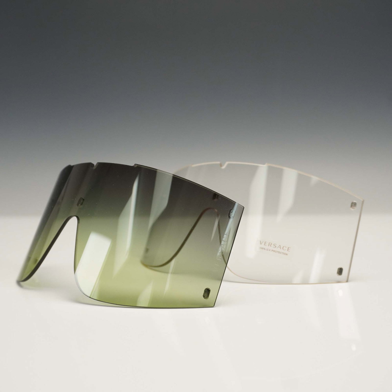 Pair of Versace VE4393 Sunglasses Shield Extra Interchangeable Lenses: Pair of Versace VE4393 interchangeable visor-style shield lenses. Includes one clear lens marked "Versace 100% U.V. Protection" and one gradient lens that transitions from dark gray to light green. Bo