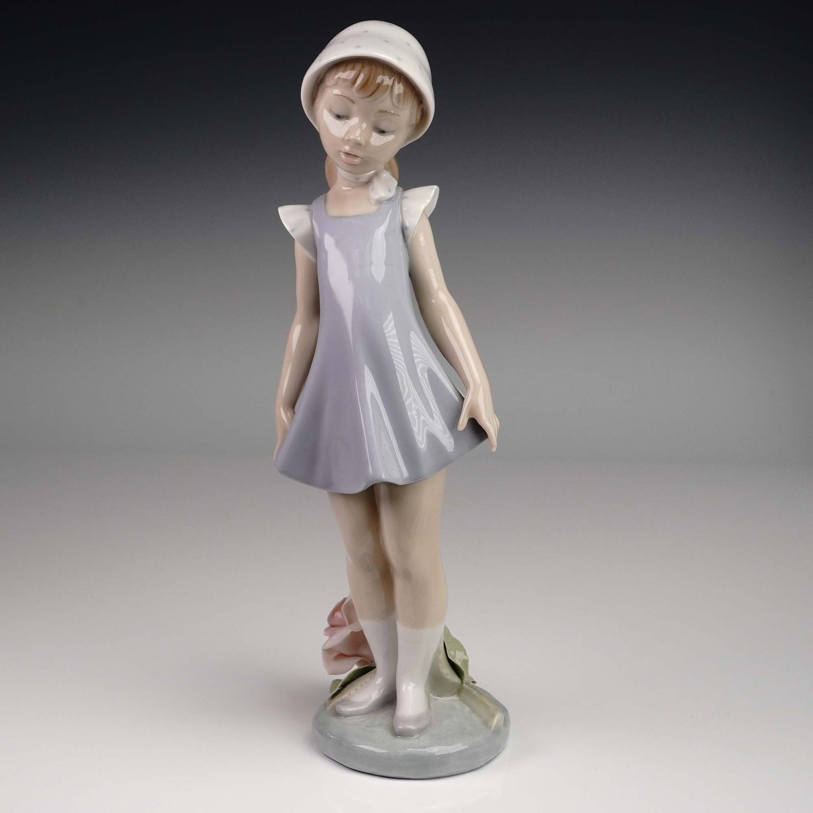 Lladro Porcelain Figurine 01005039, Candid: This Lladro porcelain figurine, model 01005039 titled Candid, features a young girl in a flared lavender dress and white bonnet, gazing demurely downward as she gently lifts the hem of her dress. A de