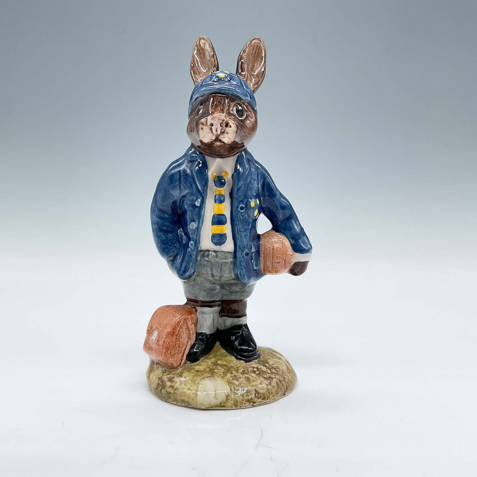 Royal Doulton Bunnykins Figurine, Schoolboy Bunnykins DB66: This Royal Doulton Bunnykins figurine, titled "Schoolboy Bunnykins," depicts a charming rabbit dressed in a blue coat, tie, and cap, carrying a satchel. Crafted in fine china, this collectible piece m