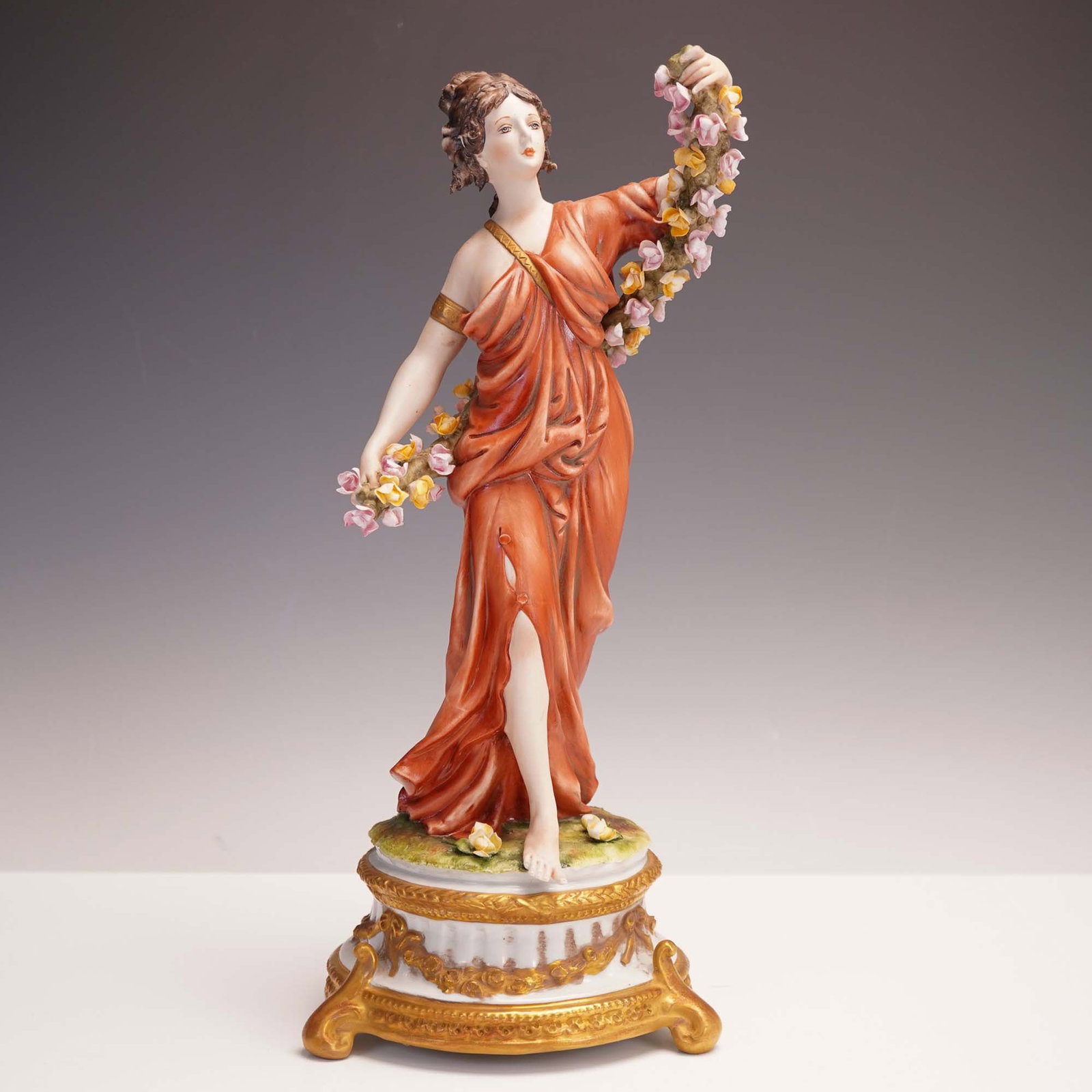 Capodimonte Porcelain Figurine, Woman with Floral Garland: An elegant Italian Capodimonte-style porcelain figurine depicting a classical woman draped in a flowing rust-colored gown, gracefully holding a garland of hand-applied flowers. Set atop a white and go