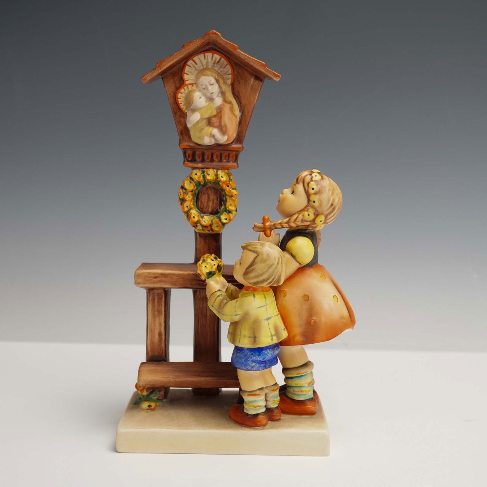 Goebel Hummel Figurine, Adoration: Charming Goebel Hummel porcelain figurine titled Adoration, featuring a young girl and boy placing a floral wreath beneath a religious shrine. Richly painted with vibrant tones and fine detail, this p