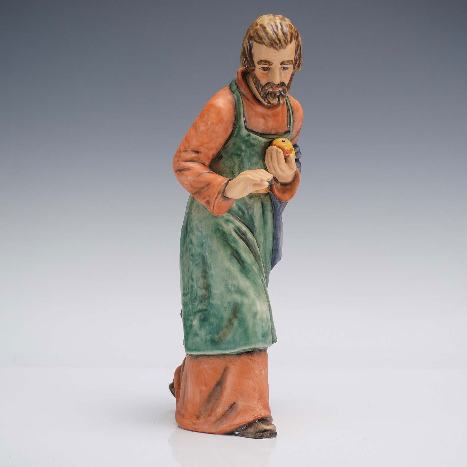 Goebel Hummel Figurine, Joseph: A finely rendered hand painted figurine depicting Joseph as part of a nativity scene, portrayed with serene humility and gentle poise. He is shown cradling two golden fruits, symbolic of sustenance an