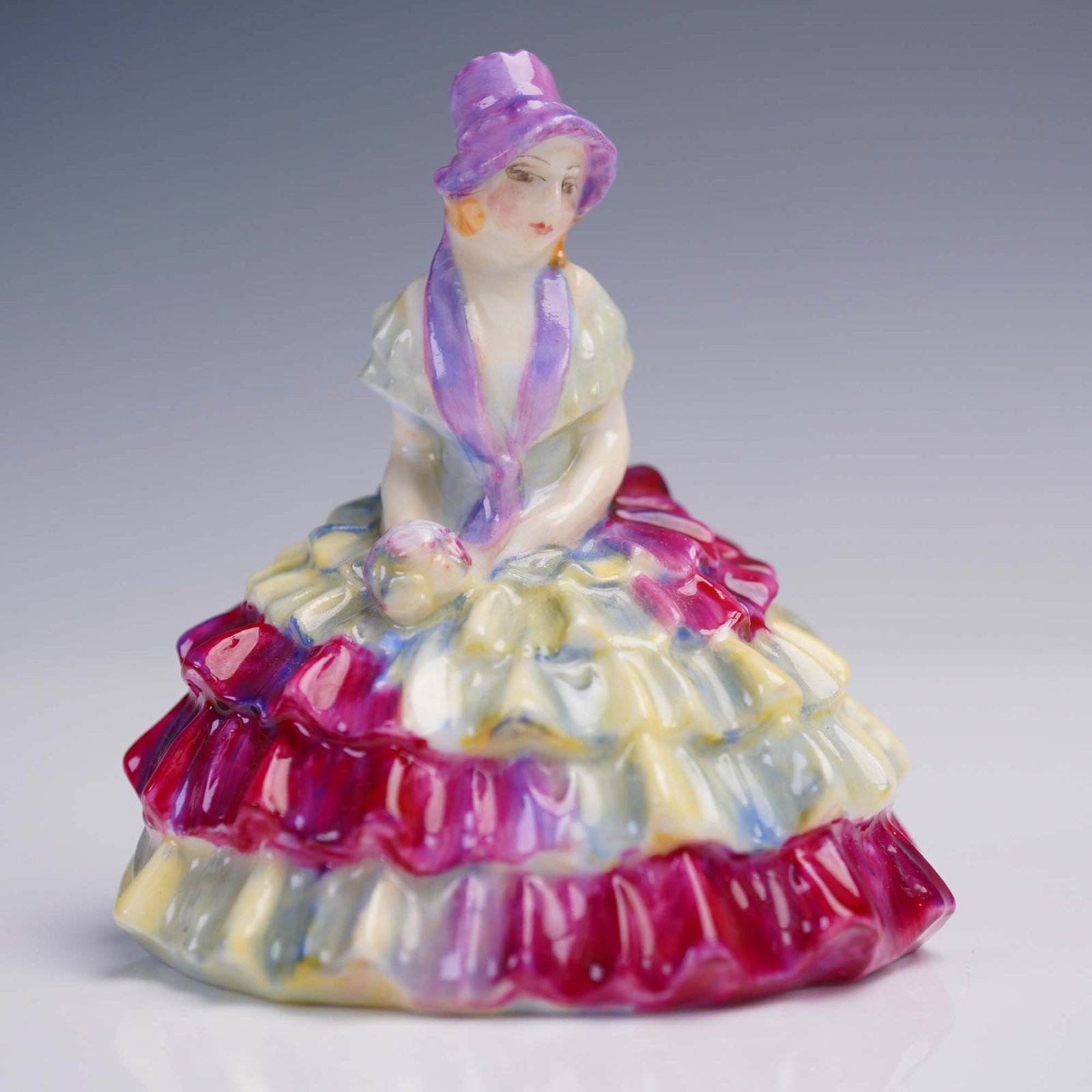 Royal Doulton Figurine, Chloe M29: This elegant Royal Doulton porcelain figurine titled Chloe features a seated young lady in a multi-colored ruffled gown with vivid tones of crimson, yellow, and lavender, and a matching bonnet. Model