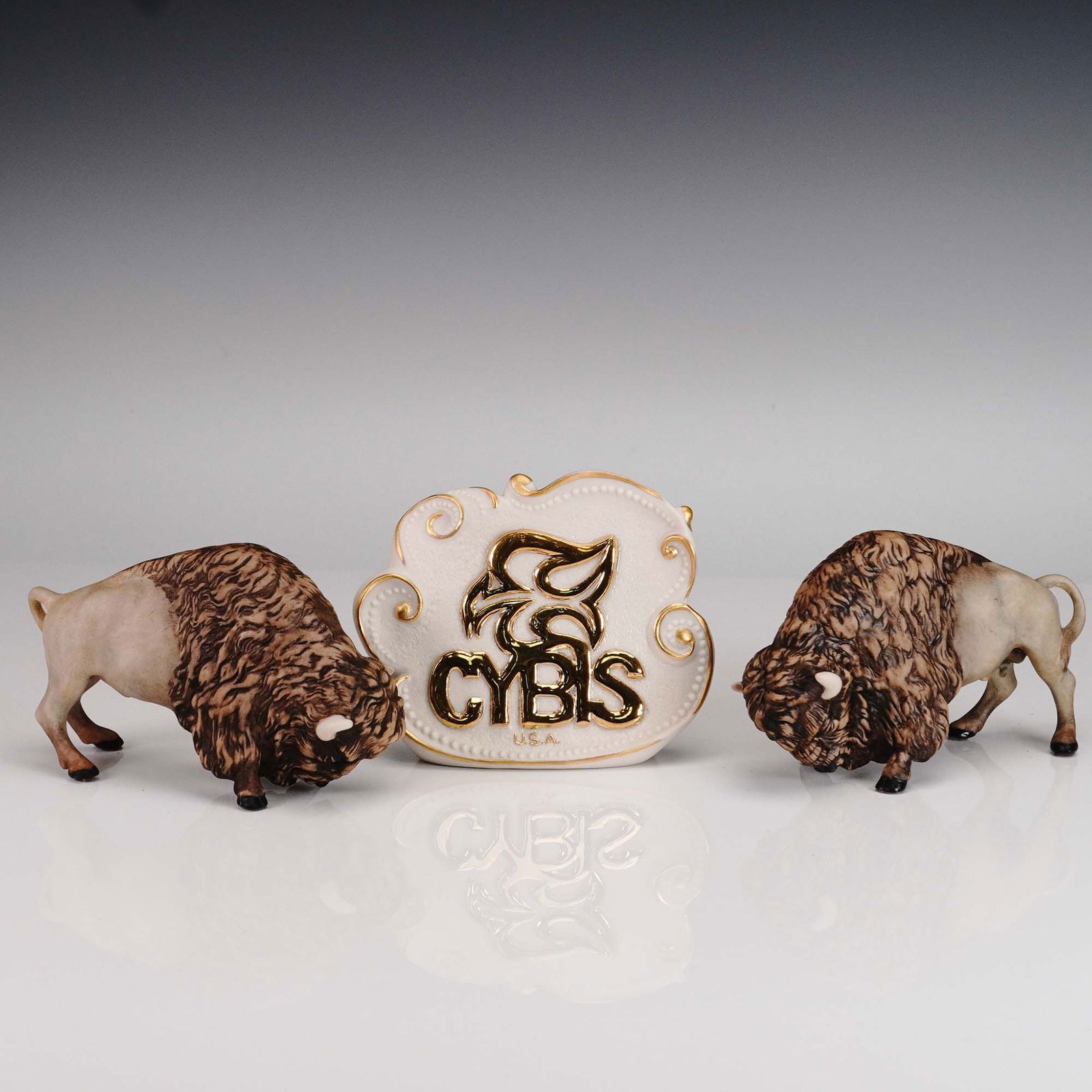 3pc Cybis Porcelain Figurines, Buffalos and Plaque: This set features two finely crafted Cybis porcelain American Bison figurines presented with a sculptural Cybis logo plaque. Each bison is rendered in naturalistic detail, showcasing the brand's hallm