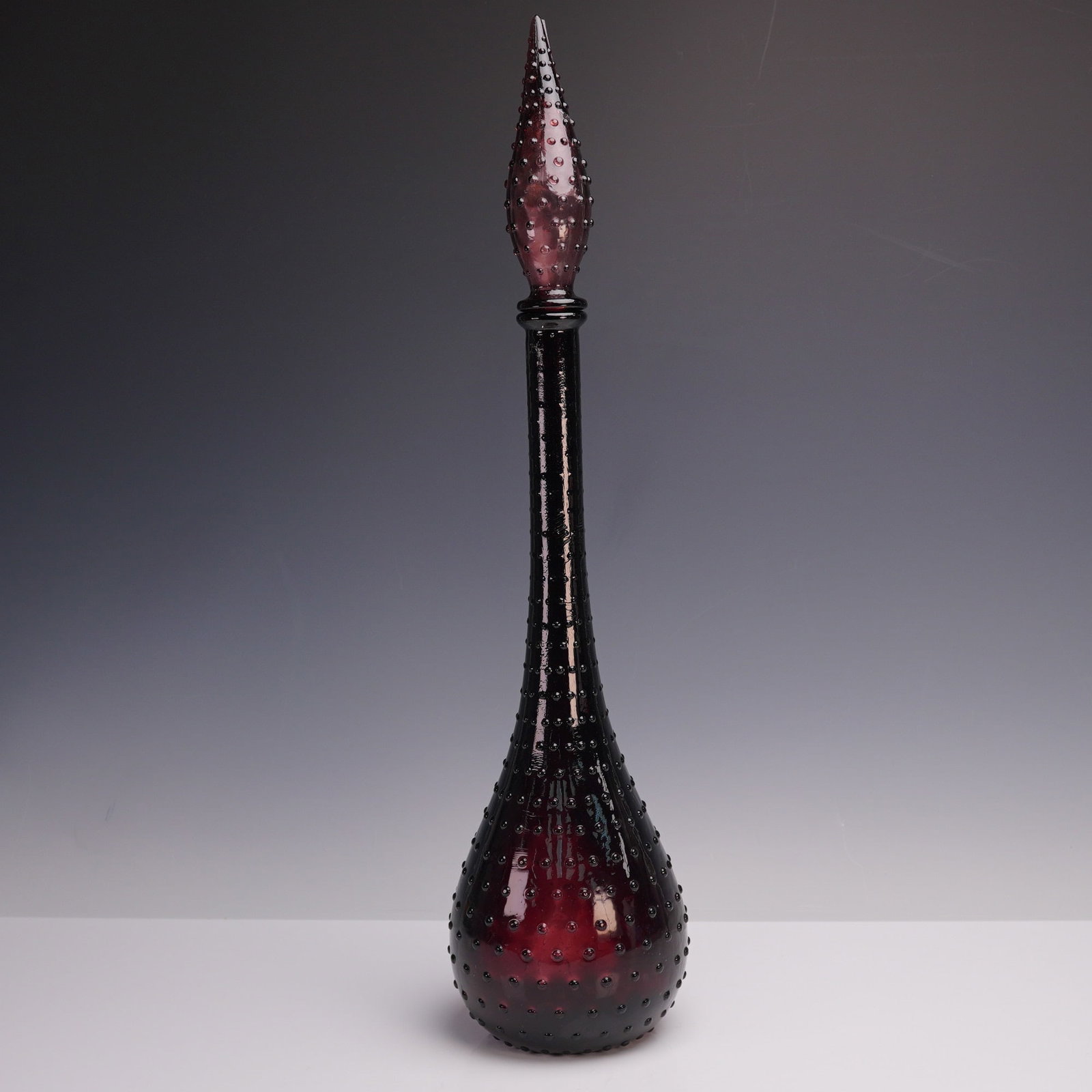 Plum Glass Empoli Bottle with Stopper: Striking in deep plum glass, this tall Empoli bottle features a classic mid century modern silhouette with a long slender neck and elegant teardrop stopper. The surface is richly textured with raised 