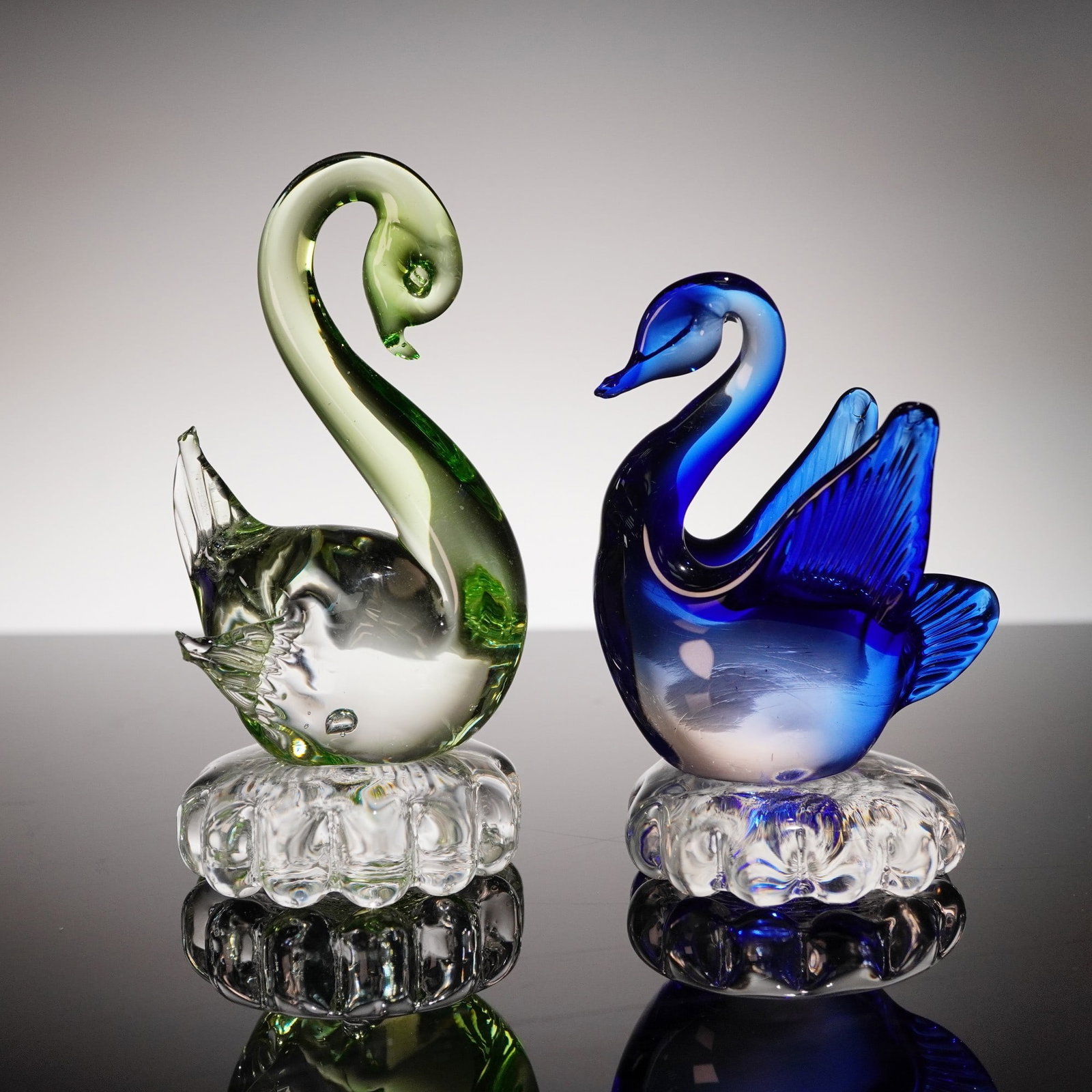 Pair of Blue and Green Glass Swan Figurines: Pair of Murano style art glass swan figurines, each sculpted with a distinctive color palette and perched atop clear glass pedestal-style bases with scalloped edges. One figurine features a deep cobal