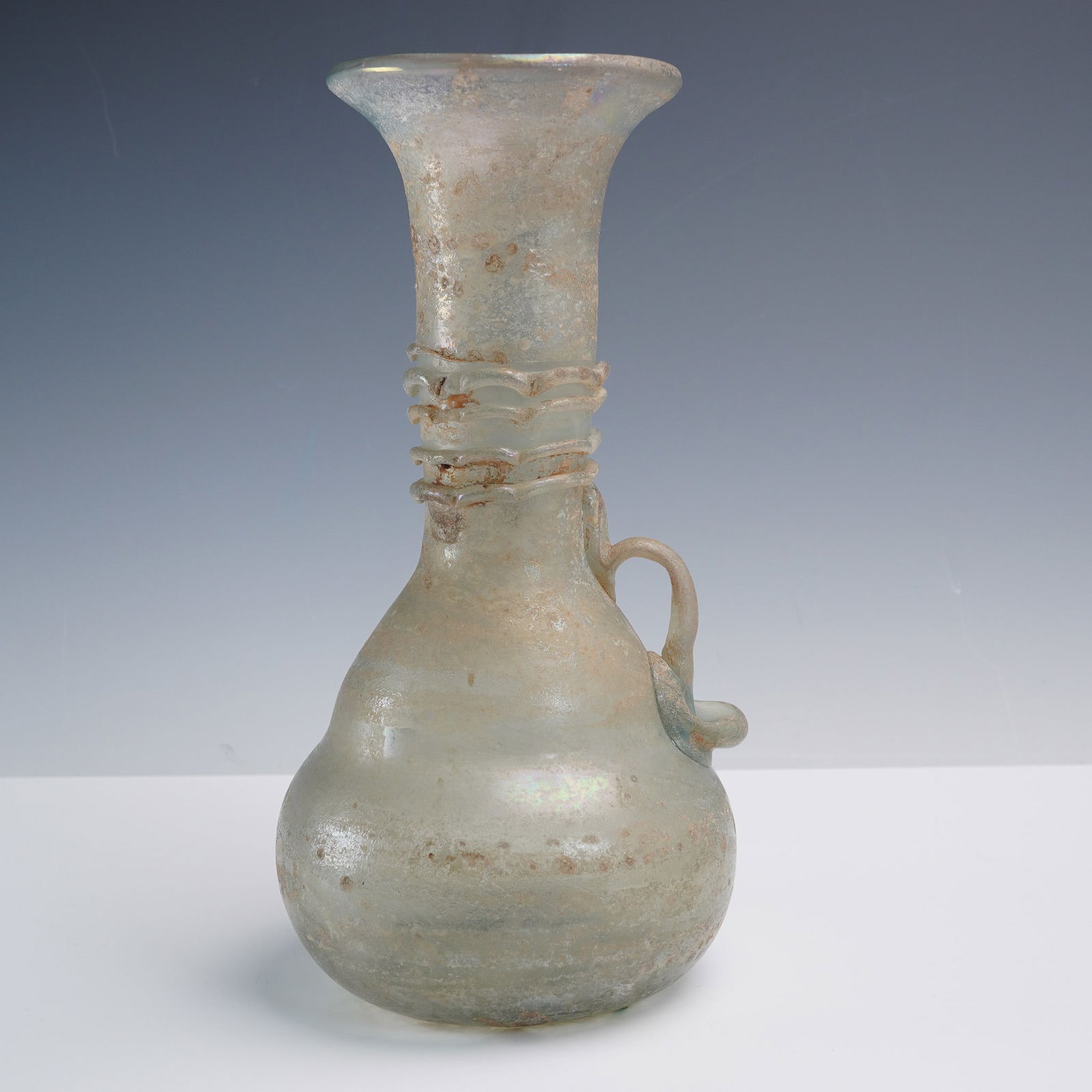 Ancient Glass Vessel with Applied Handle: This ancient-style glass vessel features a delicately rounded body, flared neck, and a single applied loop handle. Crafted in translucent glass with natural iridescence, the surface displays aged pati