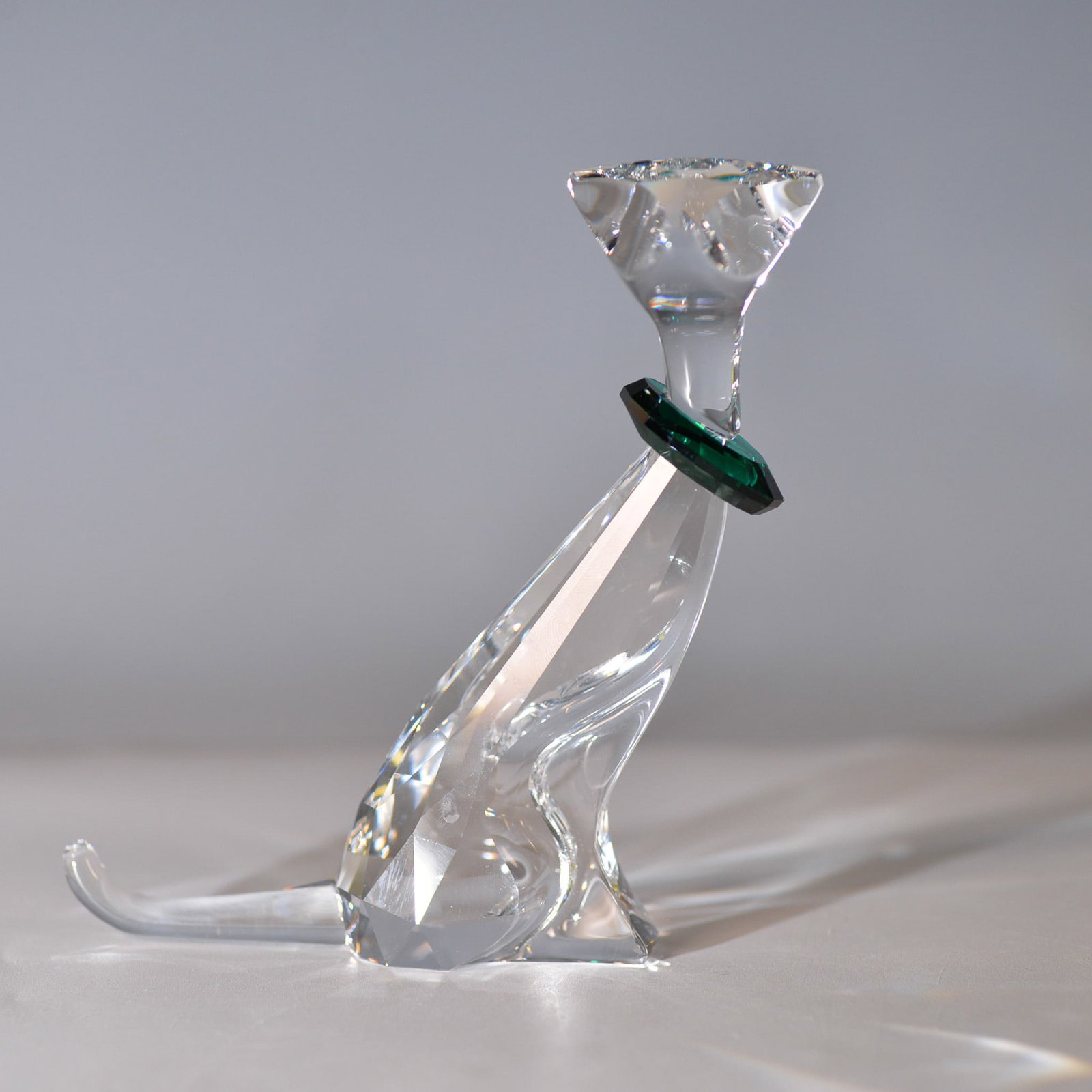 Swarovski Crystal Figurine, Symbols, the Cat: This exquisite Swarovski crystal figurine features a sitting cat adorned with a vibrant green crystal collar. The expertly crafted clear faceted body captures and reflects light beautifully, showcasin