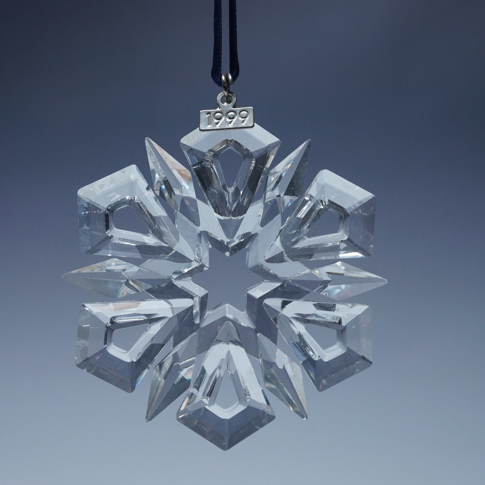 Swarovski Crystal 1999 Christmas Ornament: The intricate snowflake design features twelve radiant points, including six kite-shaped extensions, beautifully cut to reflect light with brilliance. Suspended from a dark blue ribbon, the ornament i