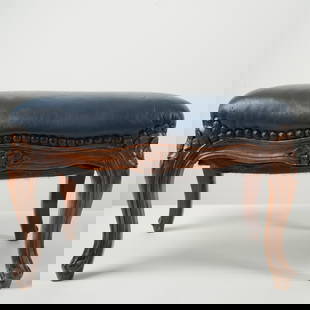 Carved Wood Ottoman with Blue Upholstered Seat: Ornately carved wooden ottoman featuring a floral motif along the apron and cabriole legs. The top is upholstered in a smooth, dark blue vinyl or leatherette with a row of decorative nailhead trim enc