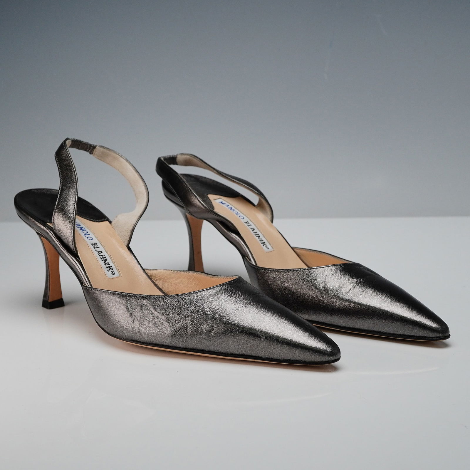 Manolo Blahnik Metallic Slingback Heels: This elegant pair of Manolo Blahnik slingback heels features a striking metallic gunmetal leather exterior with a sleek pointed toe silhouette and a mid-height stiletto heel. The interior is crafted w
