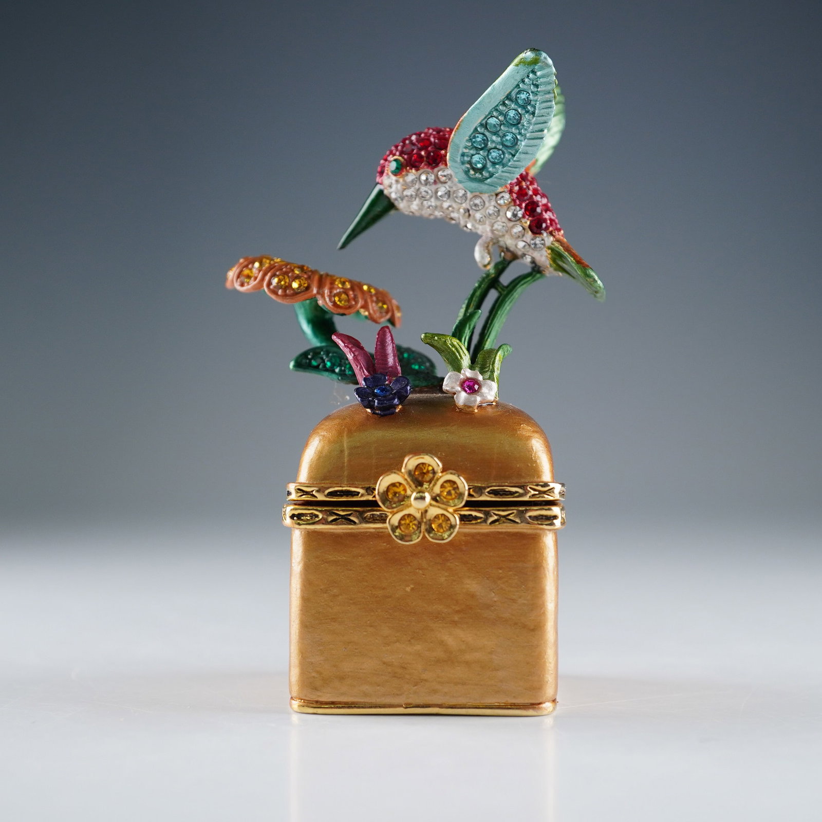 Rucinni Jeweled Hummingbird Trinket Box: Rucinni vibrant trinket box designed as a blooming flower being visited by a jeweled hummingbird, perched atop a rich copper pedestal-style base. The hinged box opens at the center of the base to reve