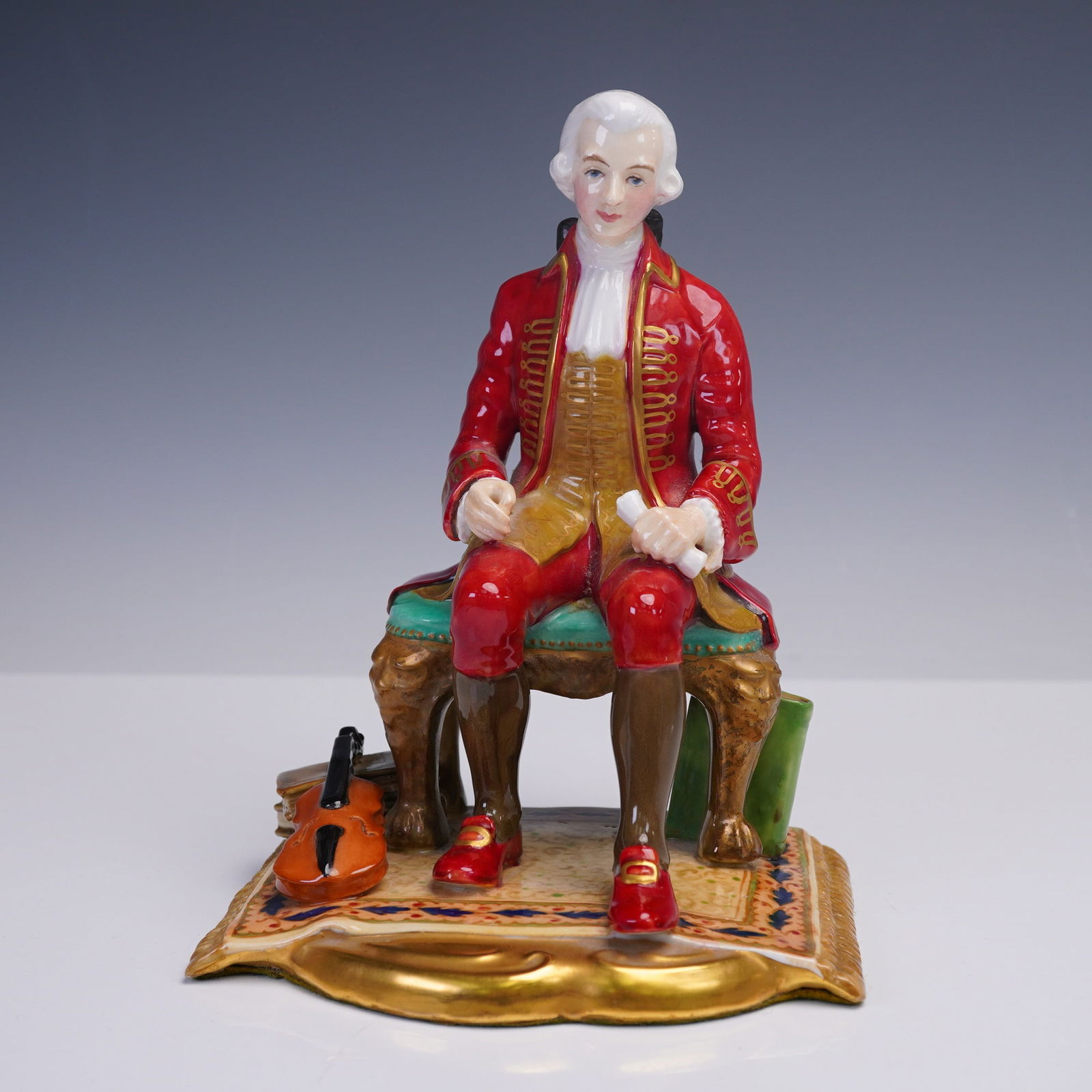 Reg Johnson Porcelain Figurine, Wolfgang Amadeus Mozart: Hand-painted porcelain figurine depicting Wolfgang Amadeus Mozart (1756 -1791), seated with a scroll in hand and a violin resting beside his foot on a stack of books. Designed by Reg Johnson under the