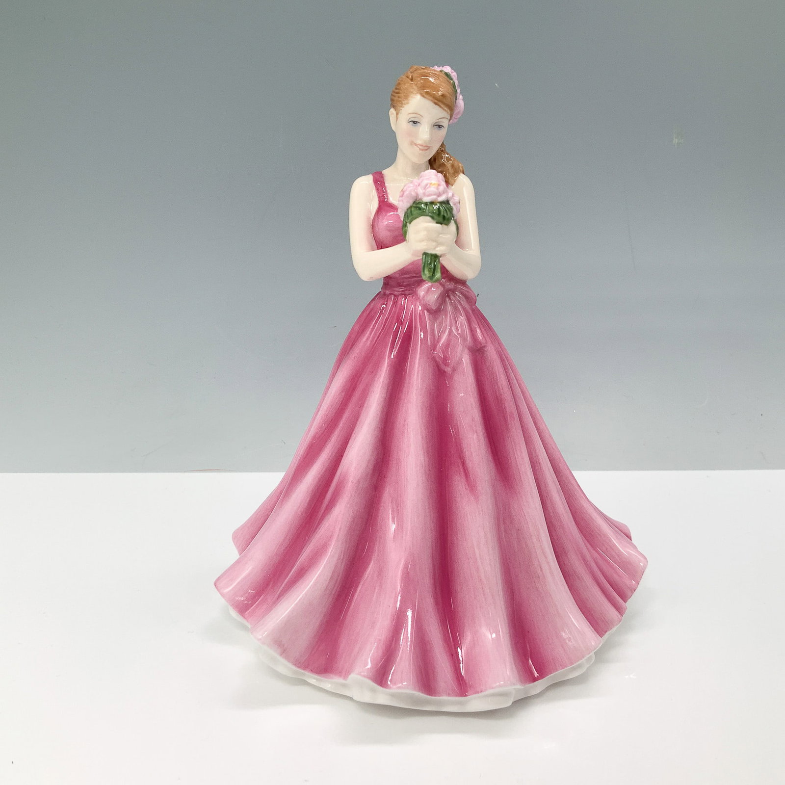 Royal Doulton Figurine, Flower of the Month May Peony HN5504: This Royal Doulton Flower of the Month series figurine, titled May Peony, features a young woman in a flowing pink gown holding a bouquet of peonies. Designed with intricate hand-painted details, this