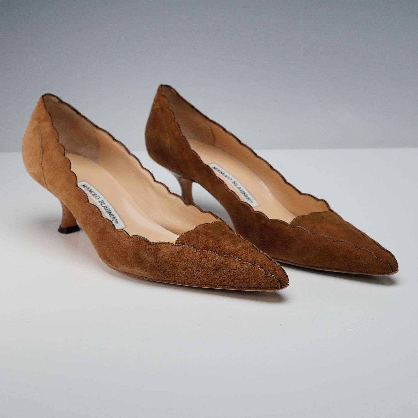 Manolo Blahnik Brown Scalloped Suede Heels: A unique and feminine pair of designer heels by Manolo Blahnik, crafted in warm brown suede with an elegant scalloped edge along the collar and vamp. The soft curves contrast beautifully with the shar