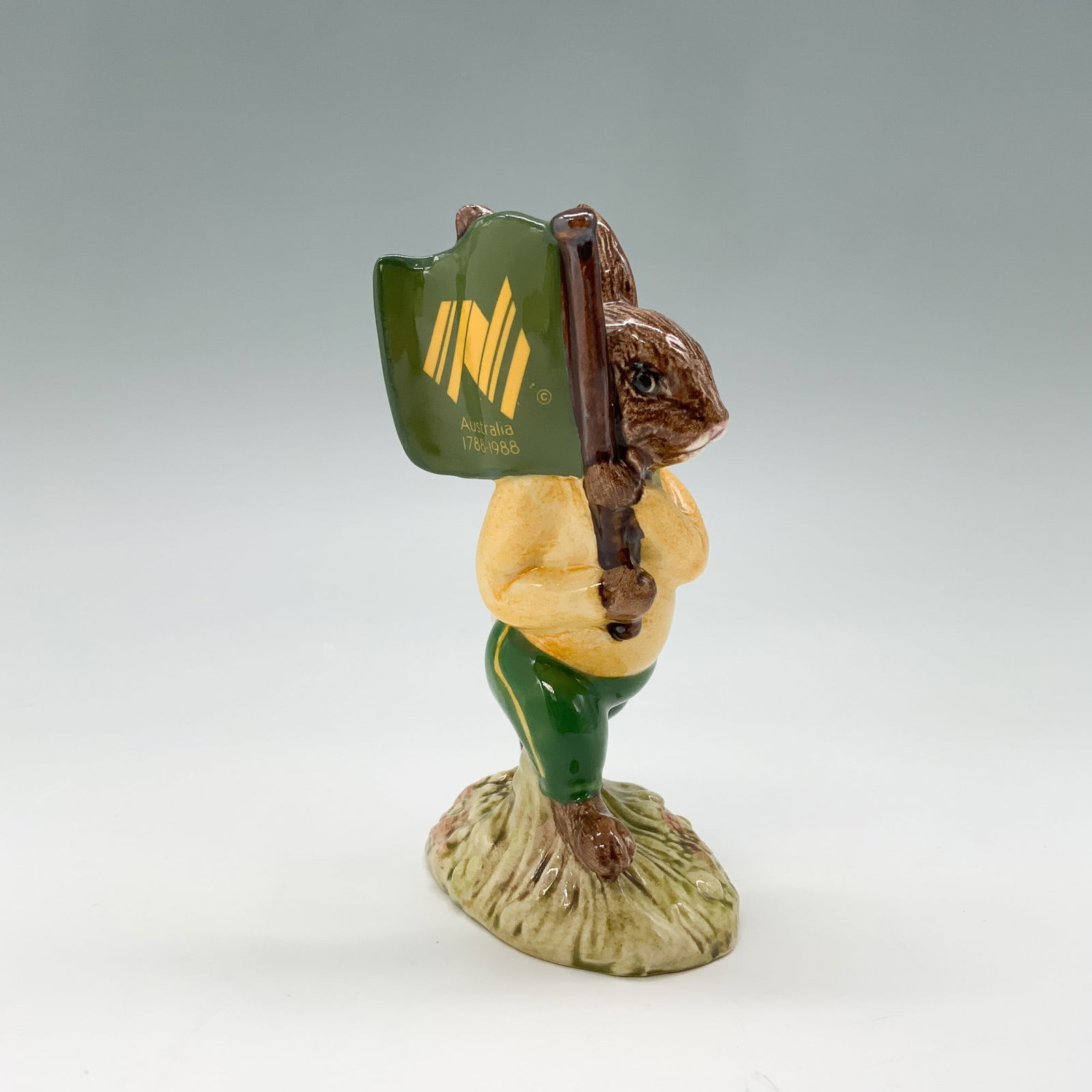 Australian Bunnykins DB58 - Royal Doulton Bunnykins: Royal Doulton Bunnykins figurine titled "Australian Bunnykins," featuring a rabbit dressed in a green and yellow tracksuit, proudly holding a green flag with "Australia 1788"1988," commemorating the A