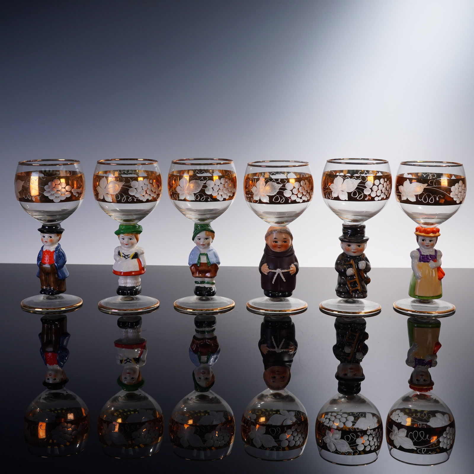 6pc West German Figural Wine Glasses with Gilt Grape Decoration: Charming set of six figural wine glasses, each standing 6 inches tall and crafted in West Germany. These whimsical glasses feature hand-painted porcelain stems sculpted in the form of various regional