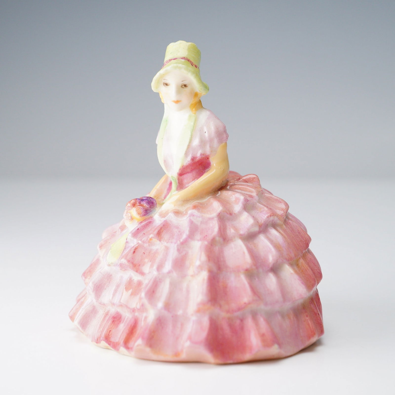 Royal Doulton Mini Figurine, Chloe M9: Miniature series. Doulton backstamp. Chloe sits patiently in her multi colored dress, pink hat and beautiful bouquet in hand. Royal Doulton backstamp. Artist: Leslie Harradine Issued: 1932 - 1945 Dime