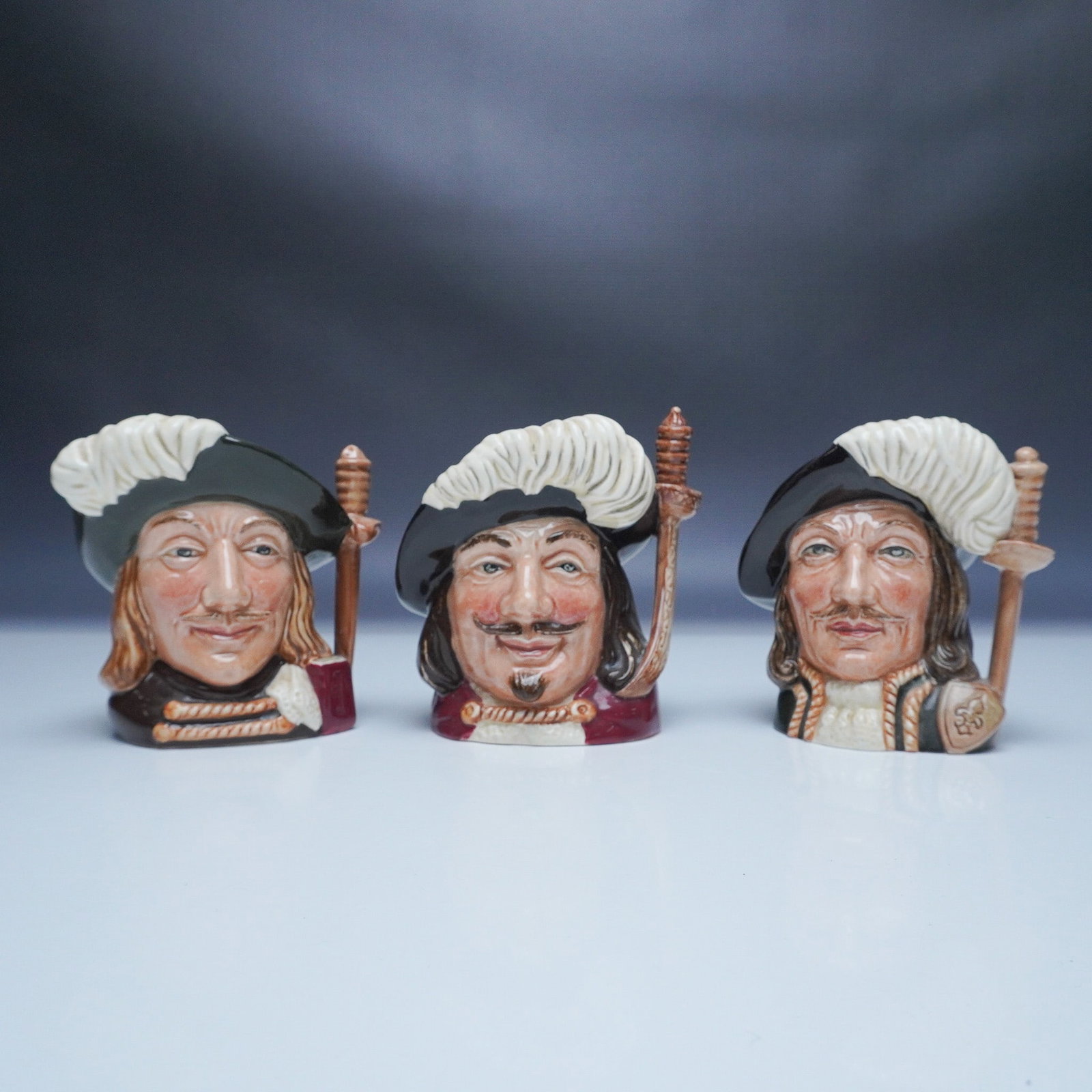 3pc Royal Doulton Character Jugs, The Three Musketeers: A set of three Royal Doulton character jugs featuring the legendary Three Musketeers: Athos (D6439), Porthos (D6440), and Aramis (D6441). Each jug is finely crafted from bone china with hand-painted d