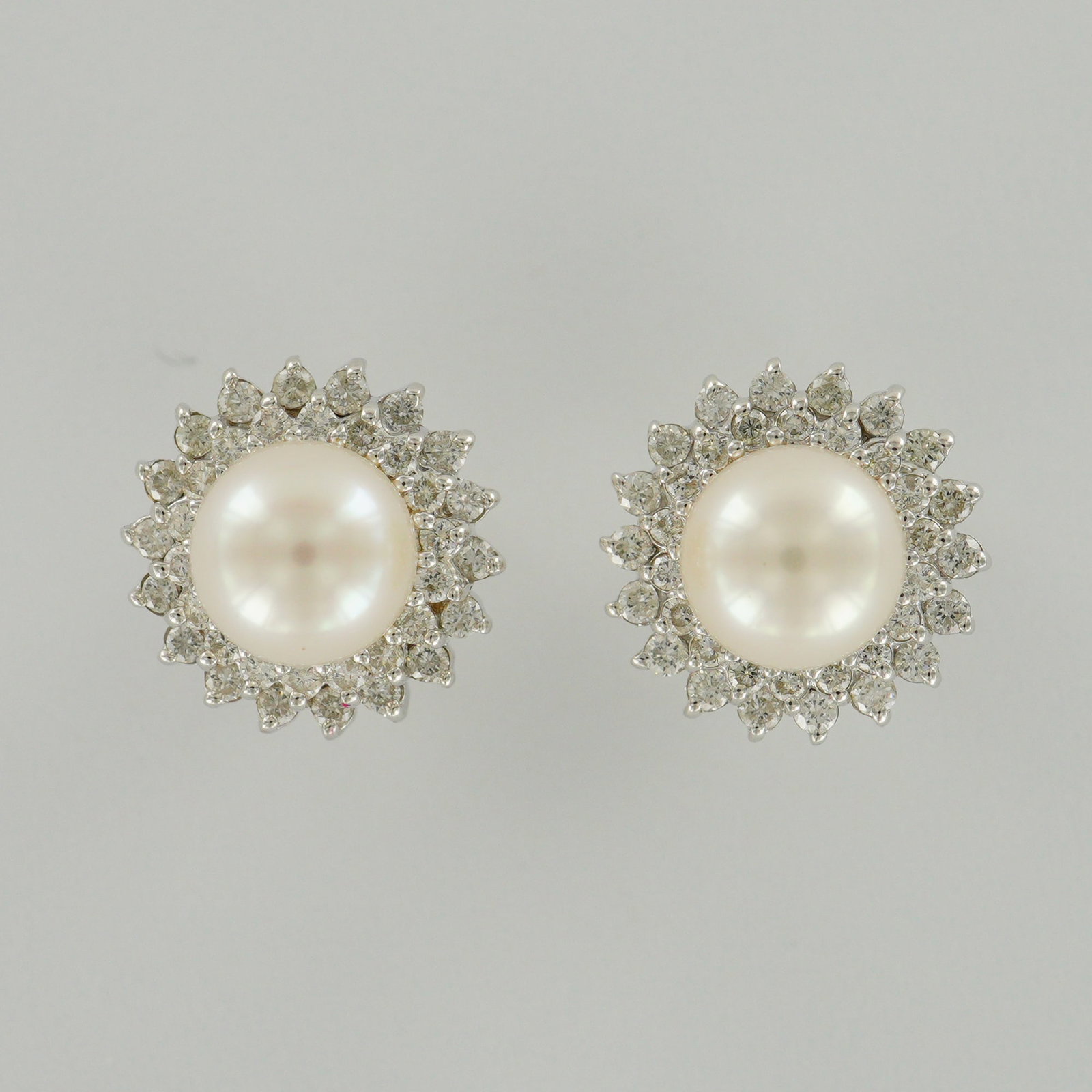 Pair of 14K White Gold Pearl and Diamond Earrings: Elegant pair of 14K white gold stud earrings featuring luminous round cultured pearls set at the center of each piece. The pearls are surrounded by two sparkling halos of brilliant-cut diamonds in a f