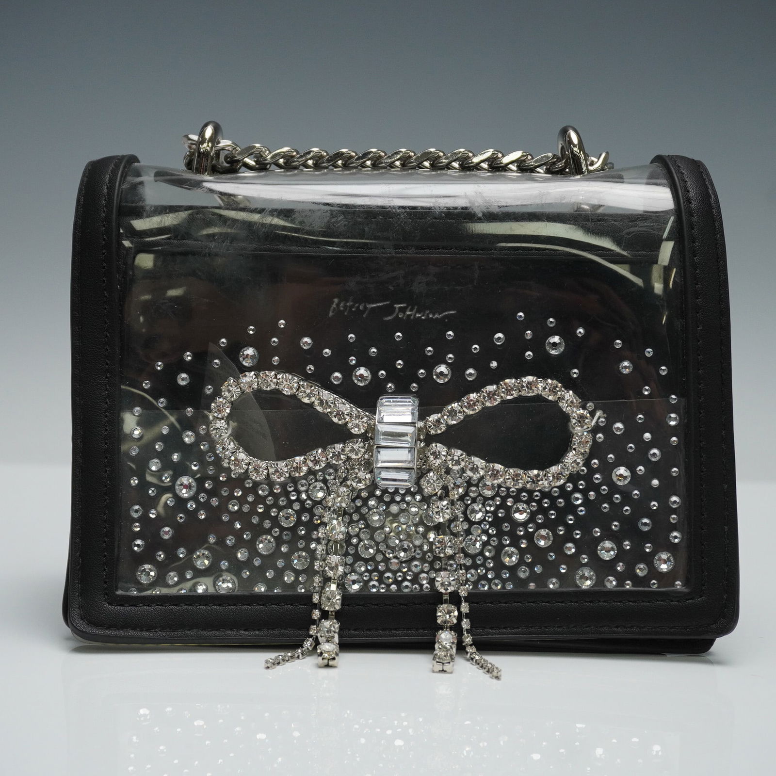 Betsey Johnson Clear Rhinestone Bow Crossbody Bag: A Betsey Johnson clear panel crossbody bag featuring a rhinestone-embellished bow design on the front. The structured form is framed with black faux leather trim and includes a silver-tone chain strap