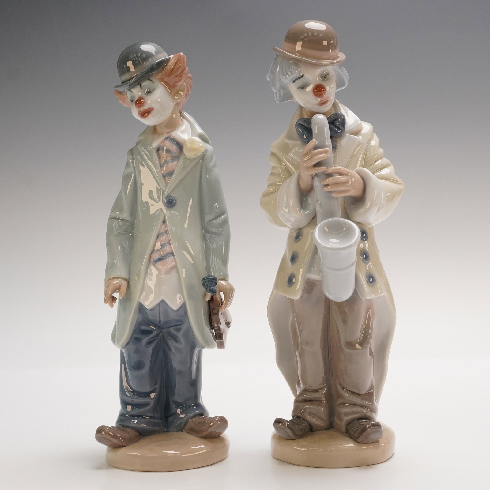 Lladro Porcelain Figurines, Circus Sam 1005472 and Sad Sax 1005471: Pair of Lladro porcelain figurines depicting expressive clowns in glossy pastel tones, including Circus Sam (1005472) holding a saxophone and Sad Sax (1005471) with a slouched posture and flower accen