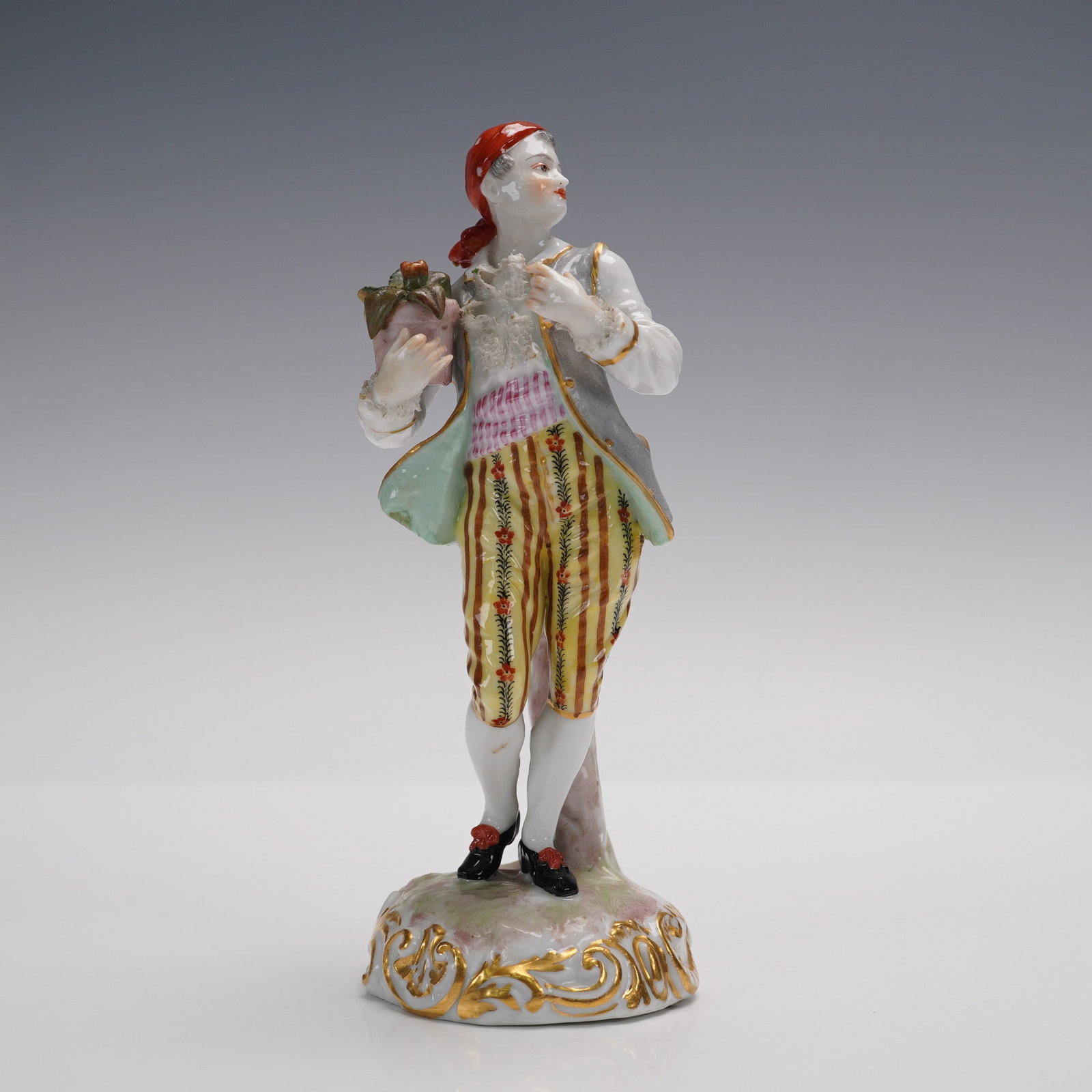 Dresden Style Porcelain Figurine, Man with Headscarf and Potted Plant: A finely painted porcelain figurine in the Dresden style depicting a young man wearing a red headscarf, striped breeches, and a gray vest, holding a potted plant in both hands. He stands beside a tree