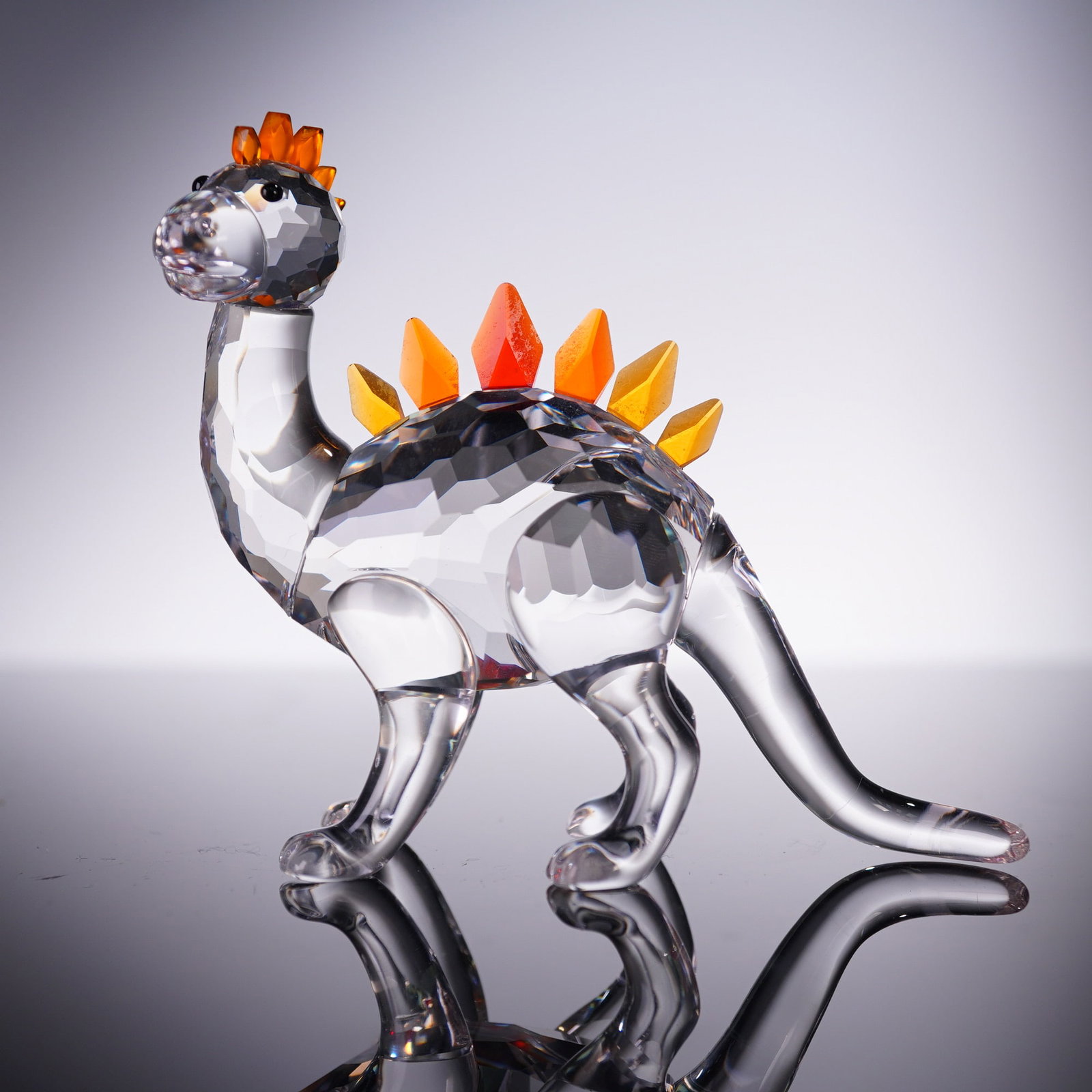Swarovski Crystal Figurine, Dino Dinosaur 268204: This whimsical crystal figurine of Dino the dinosaur model 268204 features a clear faceted body with bright red and orange crystal plates along its back. Swarovski backstamp. Includes original round b