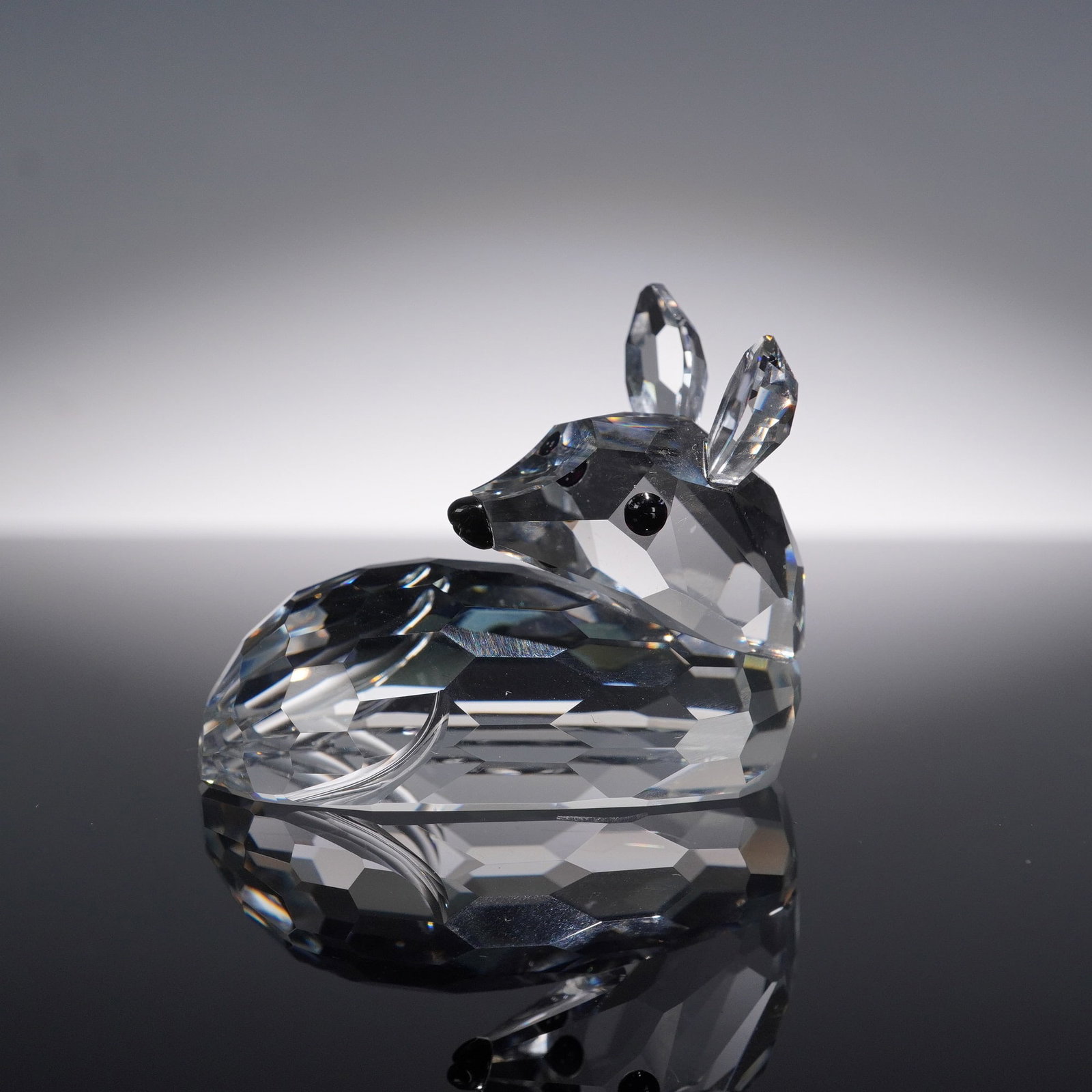 Swarovski Crystal Deer Fawn Figurine with Original Box: Swarovski crystal figurine of a resting deer fawn featuring a faceted clear body, upright ears, and black crystal eyes and nose. The smooth, compact pose and detailed facets capture the gentle charact