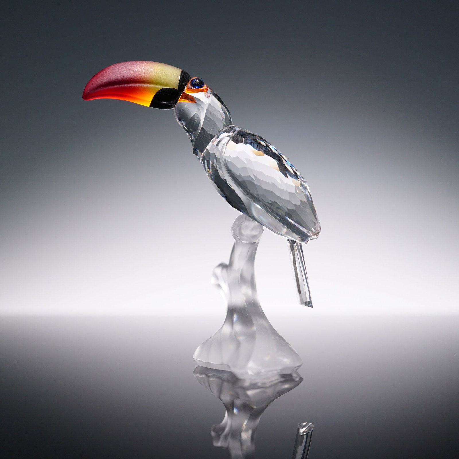 Swarovski Crystal Figurine, Toucan 234311: Vibrant Swarovski crystal figurine titled Toucan, item number 234311. Features a clear faceted crystal body perched on a frosted crystal branch, accented by a striking gradient beak in red, yellow, an