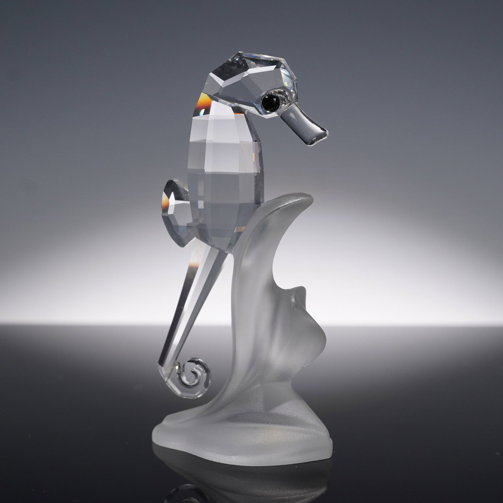 Swarovski Crystal Seahorse Figurine with Original Box: Swarovski crystal figurine of a seahorse posed upright with a faceted clear crystal body, coiled tail, and jet black eyes. The figure is mounted on a frosted crystal wave-form base, evoking the motion