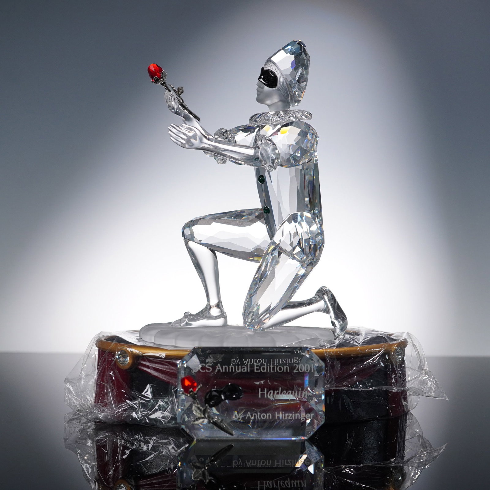 Swarovski Crystal Harlequin Figurine with Display and Plaque: 1999 Swarovski crystal Harlequin figurine issued as the SCS Annual Edition from the Masquerade trilogy, designed by Anton Hirzinger. The kneeling figure holds a red crystal rose and features expressiv