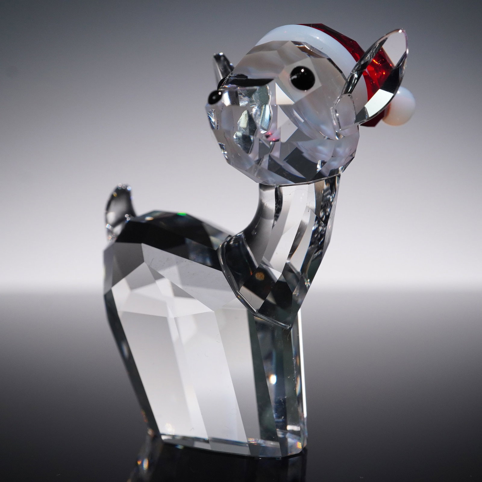 Swarovski Crystal Figurine, Doe with Santa's Hat, 5135853: Charming Swarovski crystal figurine titled Doe with Santa's Hat, item number 5135853. This clear crystal doe features a festive red-and-white Santa hat with striped detail and jet black eyes. Etched w