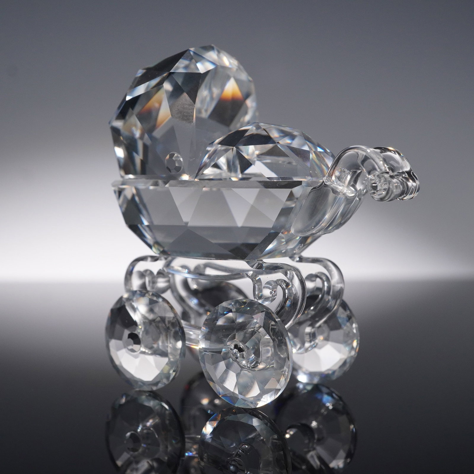 Swarovski Crystal Baby Carriage Figurine with Original Box: Swarovski crystal figurine of a baby carriage featuring a faceted bassinet design with raised hood and rounded body, set atop four clear crystal wheels with detailed scrollwork. The high level of cut 
