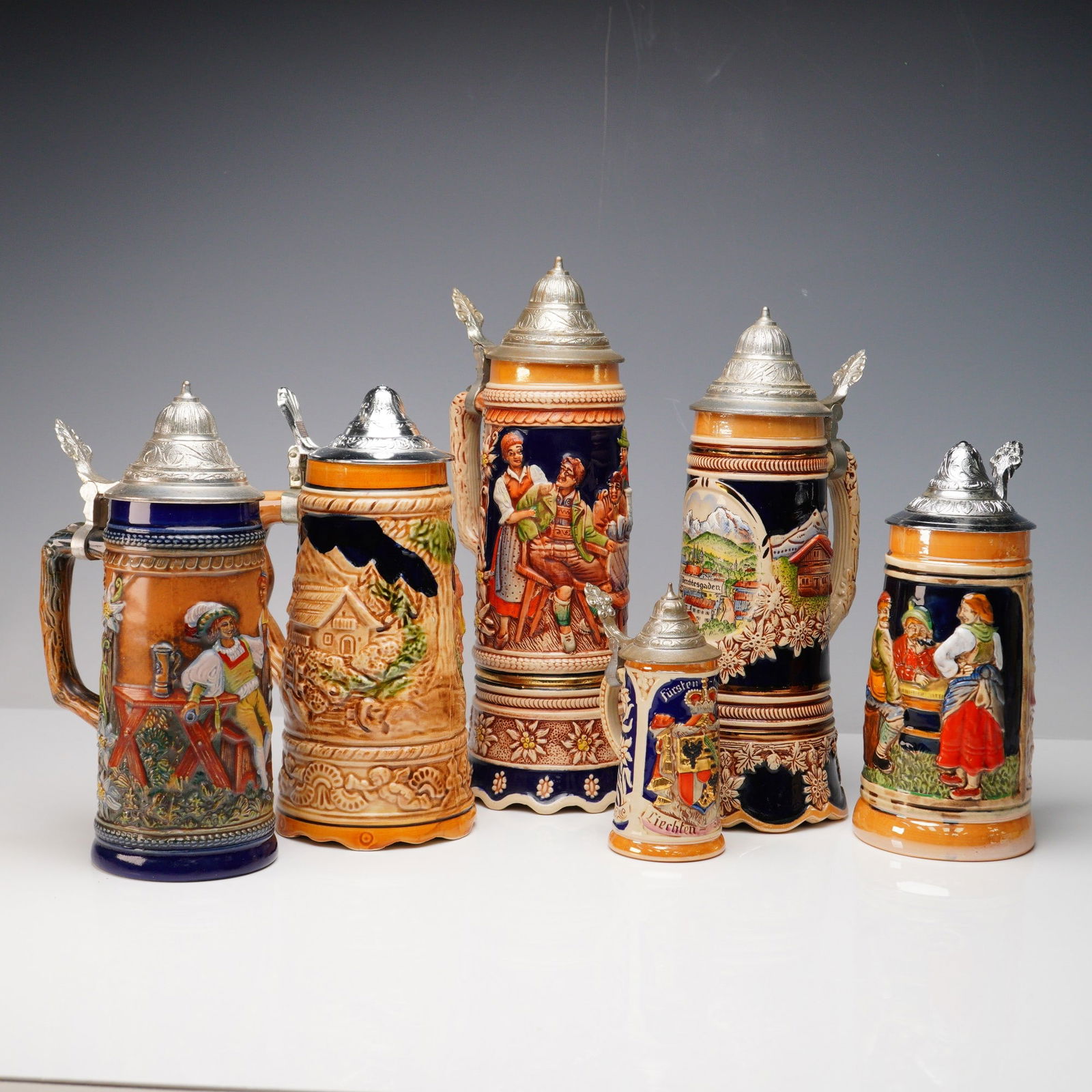 6pc German Beer Steins with Metal Lids Plus Music Box: Collection of six German beer steins featuring traditional figural and scenic relief motifs, each topped with decorative pewter lids. The group includes five standard steins with richly colored glazes