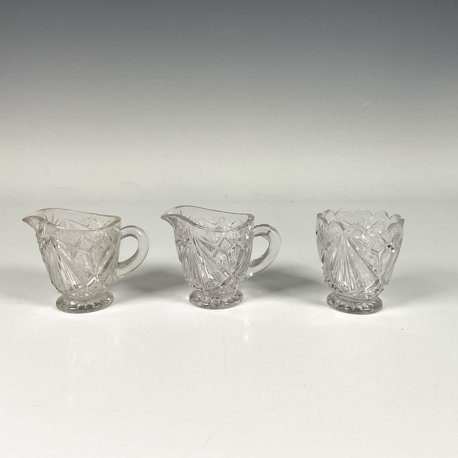 3pc Glass Toy Creamers and Child's Spooner: Two glass creamers and a child spooner in fancy cut style. Creamer: 3"L x 2"W x 2.5"H. Spooner: 2"W x 2.5"H. Dimensions: See Description