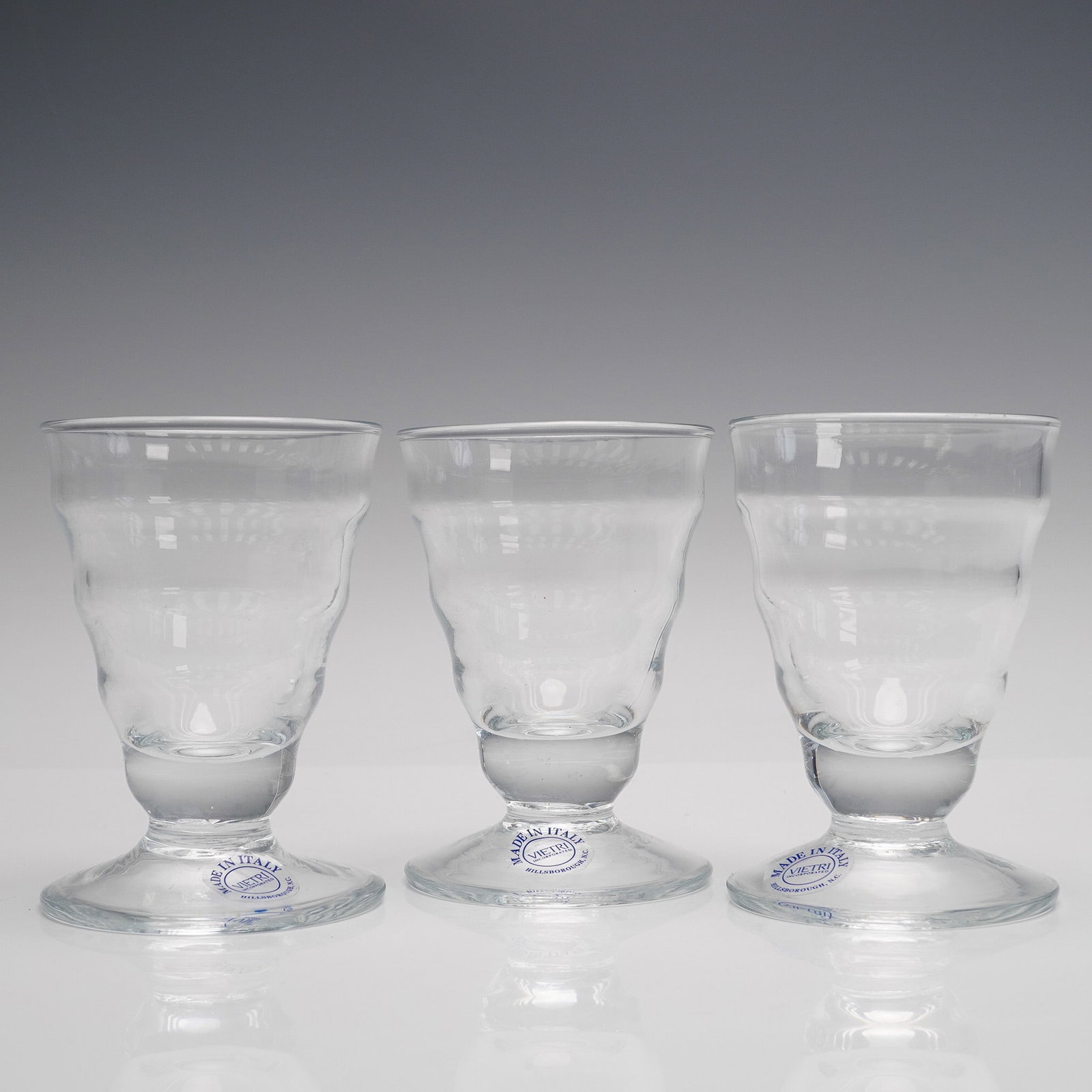 3pc Vietri Clear Glass Footed Tumblers: A set of three footed tumblers by Vietri a brand renowned for its Italian artistry and craftsmanship. These glasses are marked on the base with a circular sticker reading made in Italy Vietri incorpor